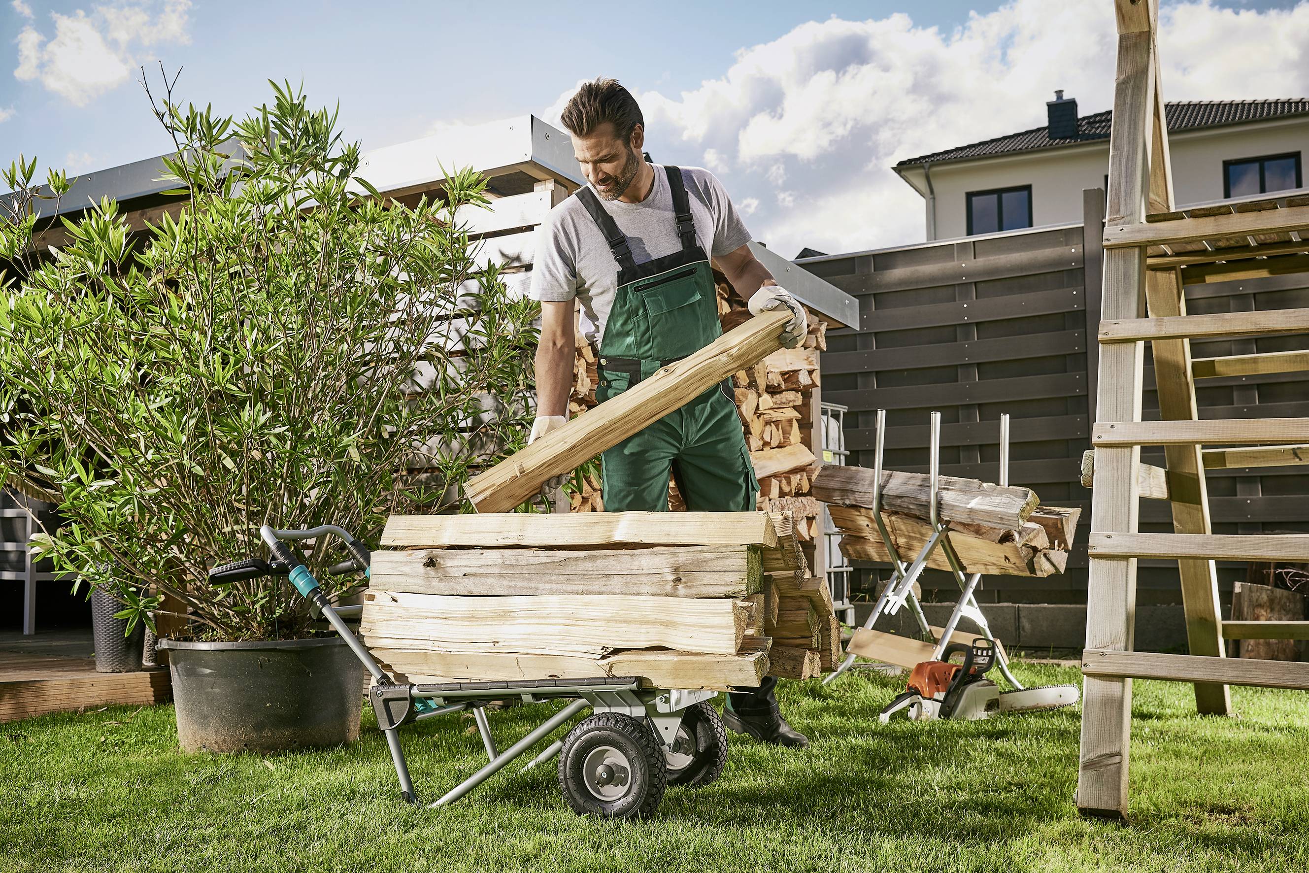 Ein Mann in Arbeitskleidung lädt in einem Garten Holzscheite in eine Schubkarre. Im Hintergrund sind ein Haus und Pflanzen sichtbar.