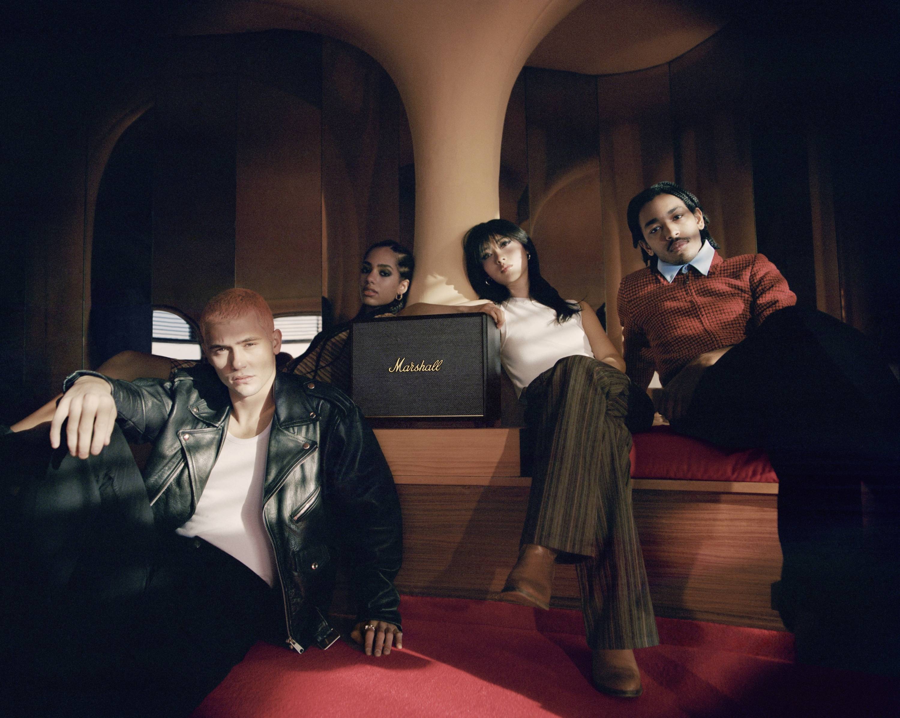 Vier Personen sitzen in einem stilvollen Raum um einen Marshall-Lautsprecher, entspannt und in modischer Kleidung.