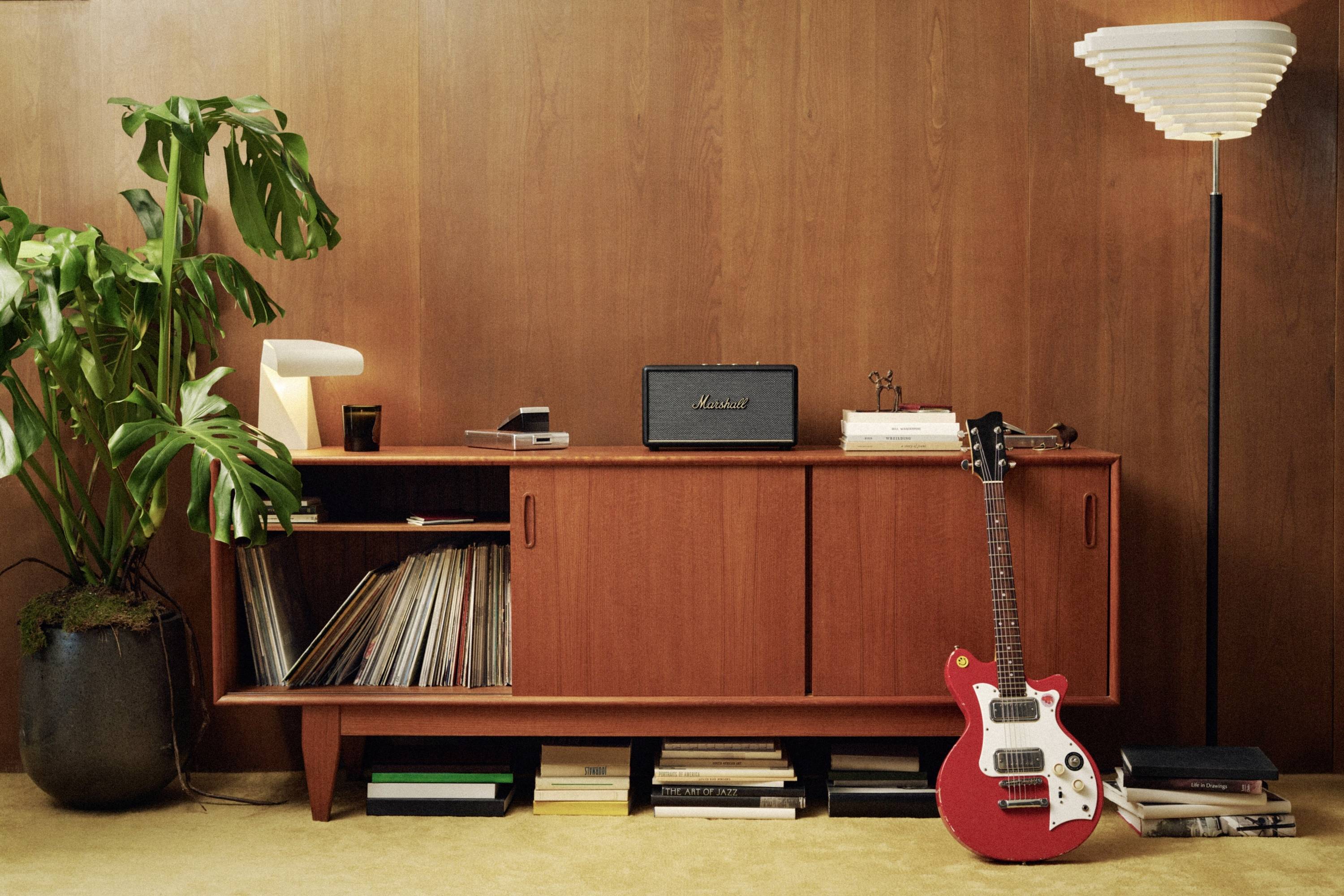 Ein rotes E-Gitarre lehnt an einem Retro-Holzschrank, der mit Schallplatten und einem Verstärker bestückt ist. Daneben eine Pflanze und Stehlampe.