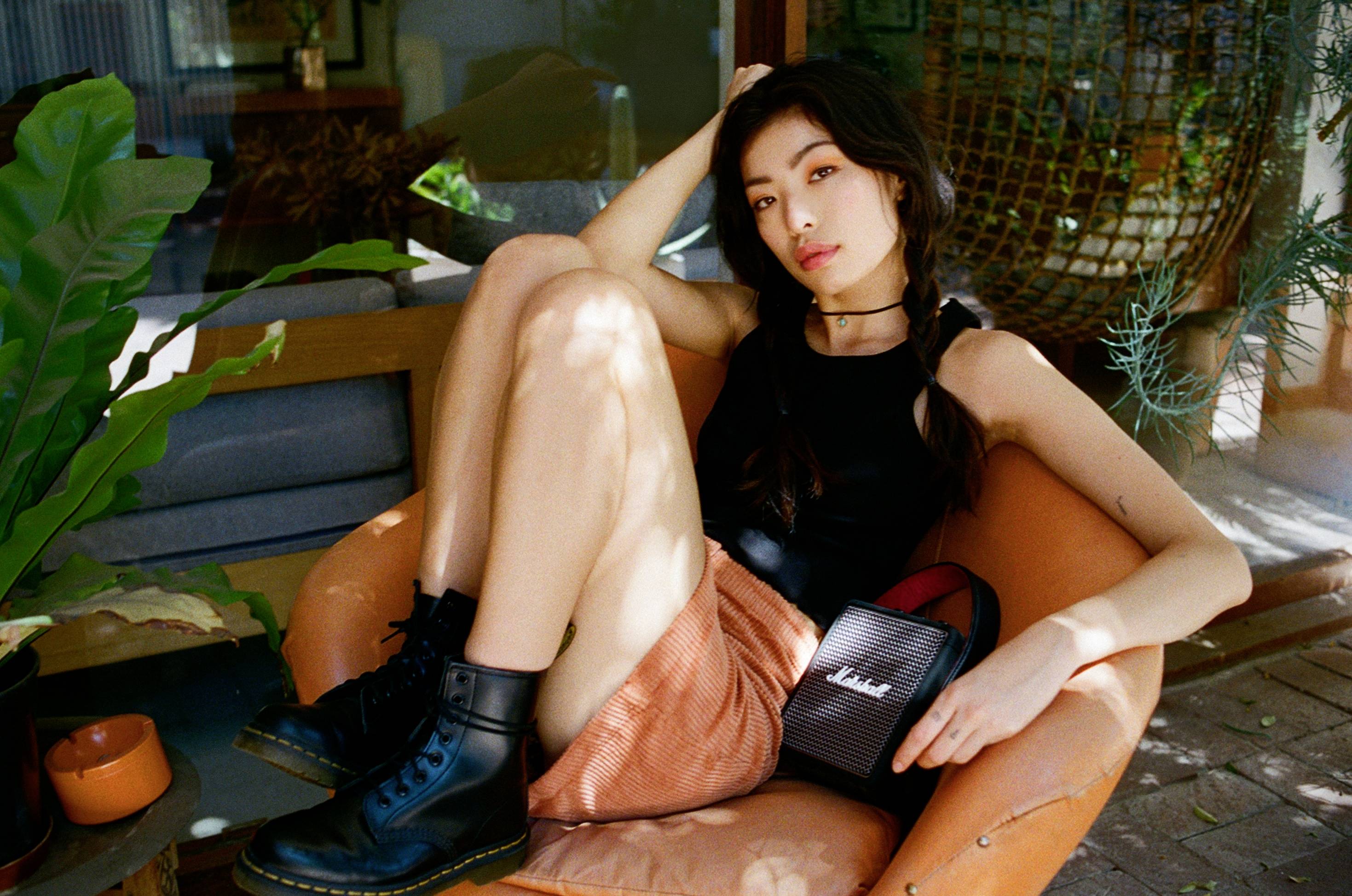 Eine Person sitzt entspannt auf einem Sessel, mit dem Arm auf der Rückenlehne. Sie trägt ein schwarzes Top, einen rosafarbenen Rock und Stiefel. Eine kleine Tasche liegt daneben.