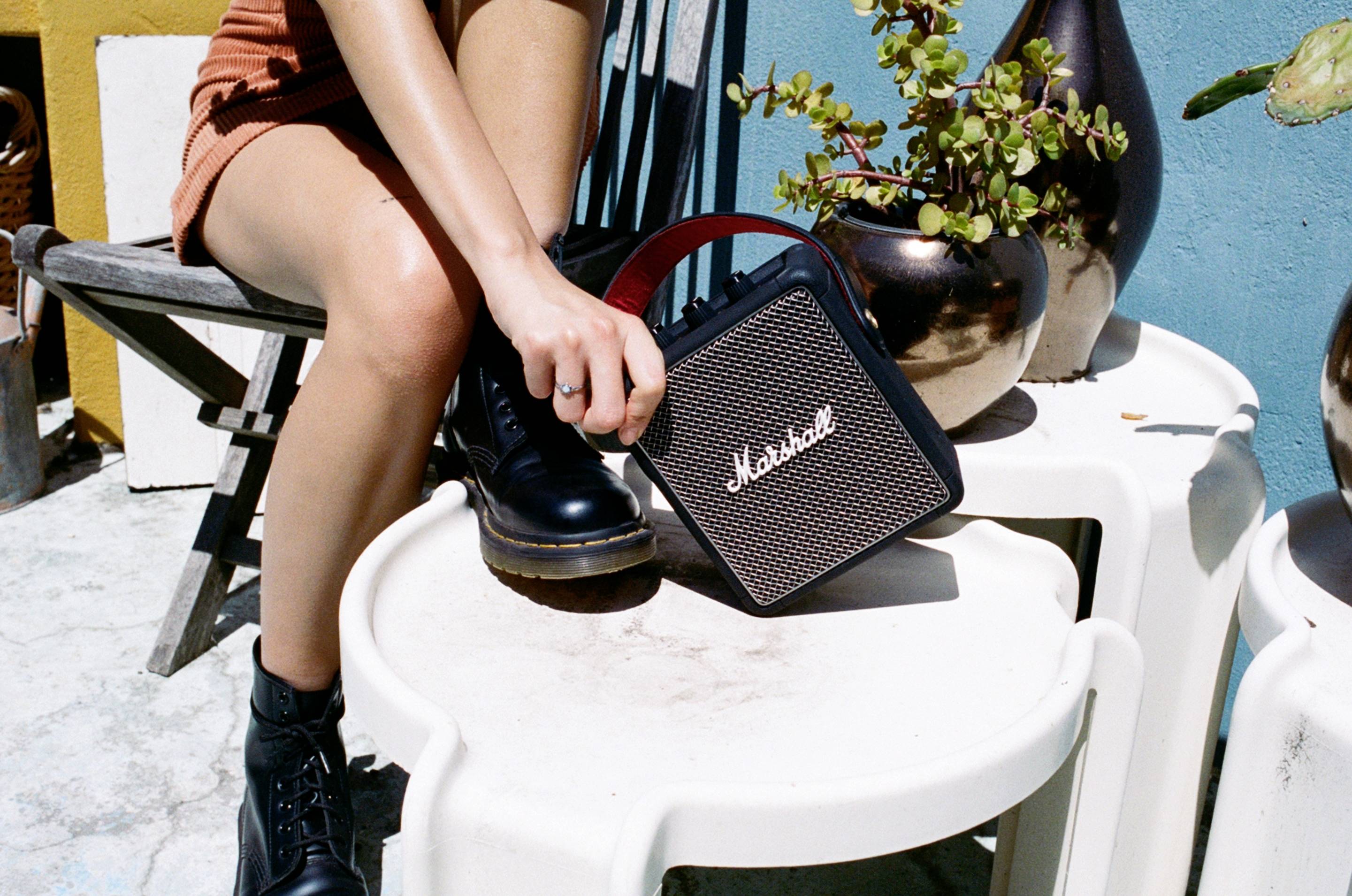 Eine Person in schwarzen Stiefeln greift nach einem tragbaren Lautsprecher auf einem Tisch im Freien. Im Hintergrund sind Pflanzen sichtbar.