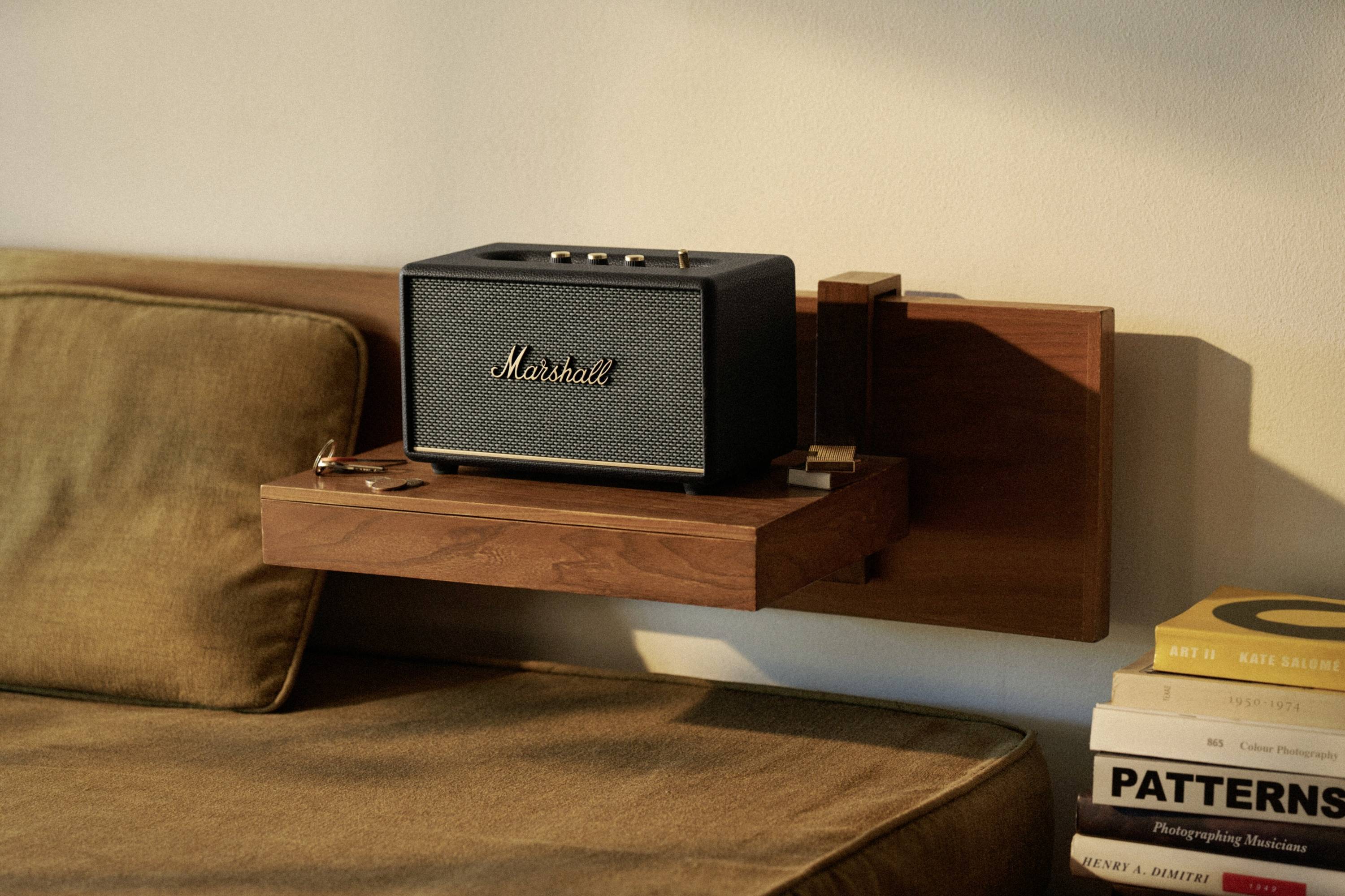 Ein schwarzer Lautsprecher der Marke 'Marshall' steht auf einem hölzernen Regal neben einem Sofa. Daneben liegen Bücher.