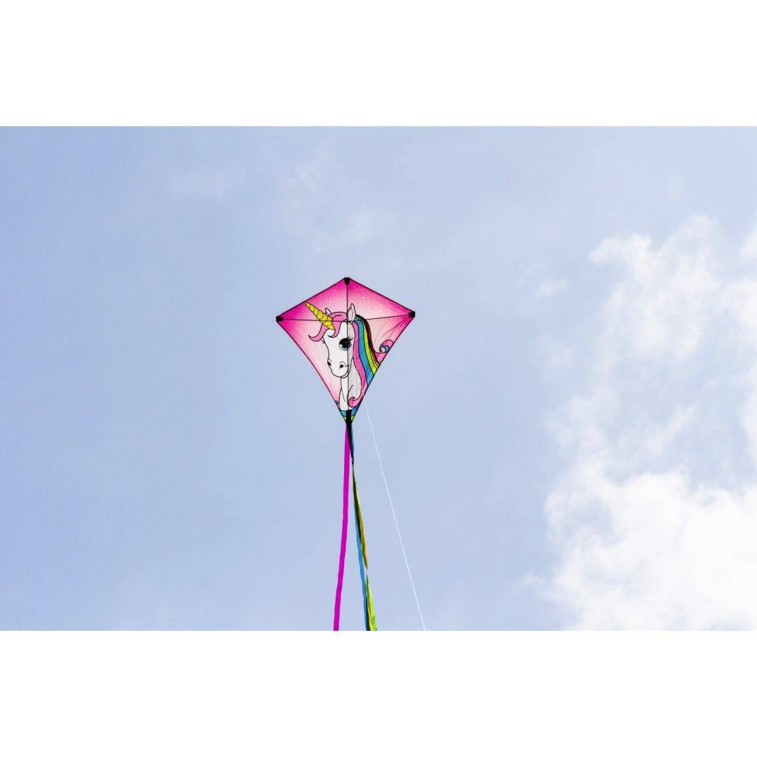 Ein rosa Drachen mit Einhornmotiv schwebt am blauen Himmel. Bunte Bänder hängen vom unteren Teil des Drachens.