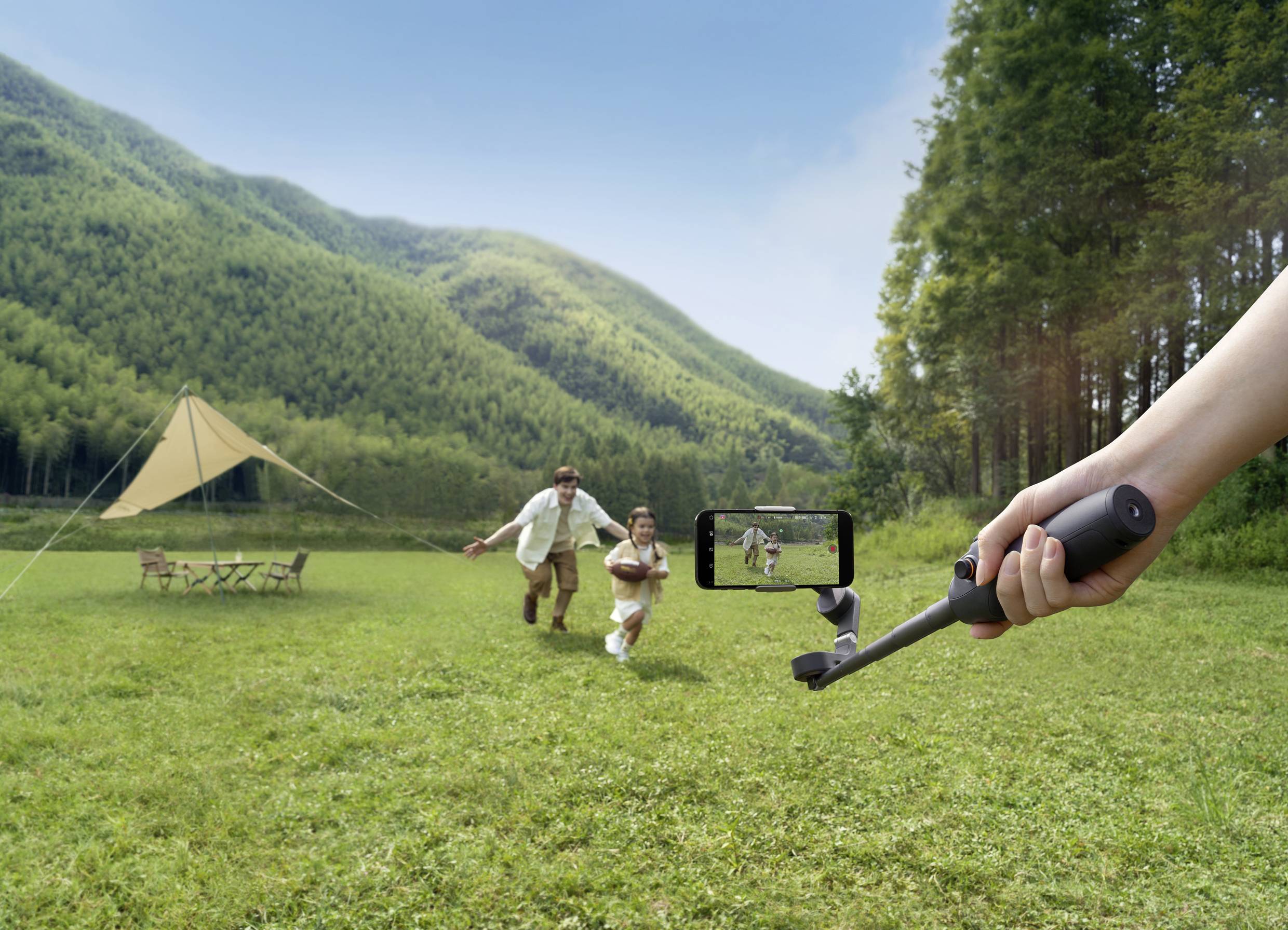 Ein Mann und ein Kind laufen im Gras in Richtung Kamera. Im Vordergrund hält eine Person ein Handy auf einem Gimbal. Im Hintergrund sind Zelte und Bäume zu sehen.