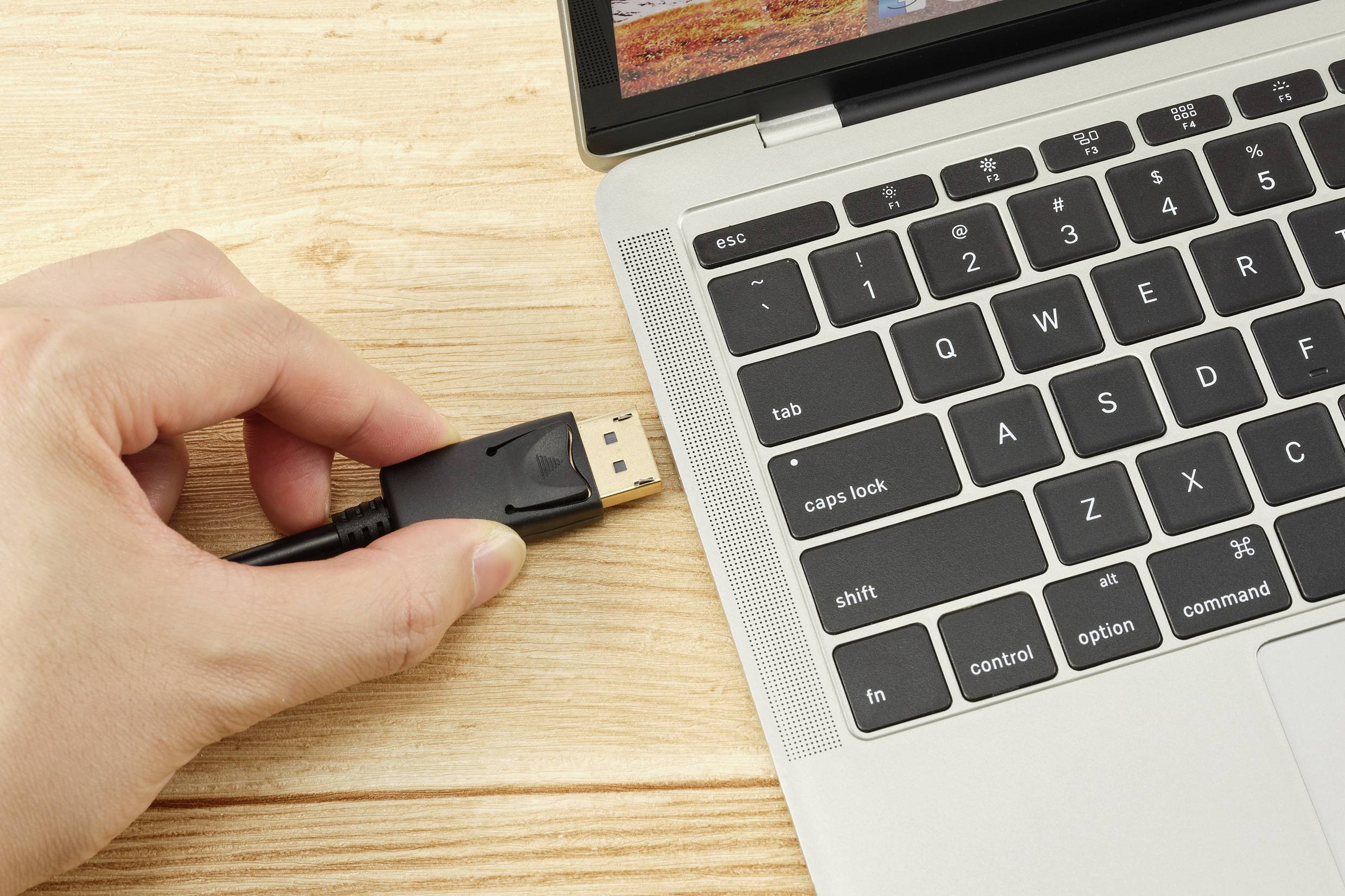 Eine Person steckt ein HDMI-Kabel in einen Laptop ein, auf einem Holztisch. Der Laptop ist geöffnet und zeigt die Tastatur.