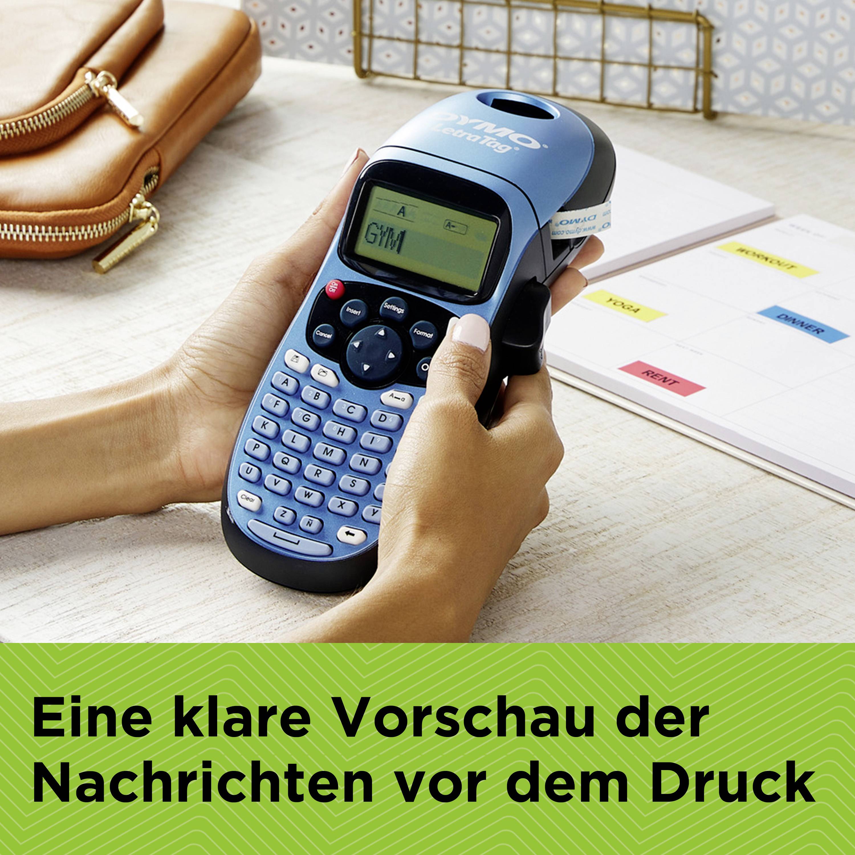 Eine Person hält ein Beschriftungsgerät, auf dem 'GM' angezeigt wird. Im Hintergrund liegt eine braune Tasche und ein Kalender auf dem Tisch.