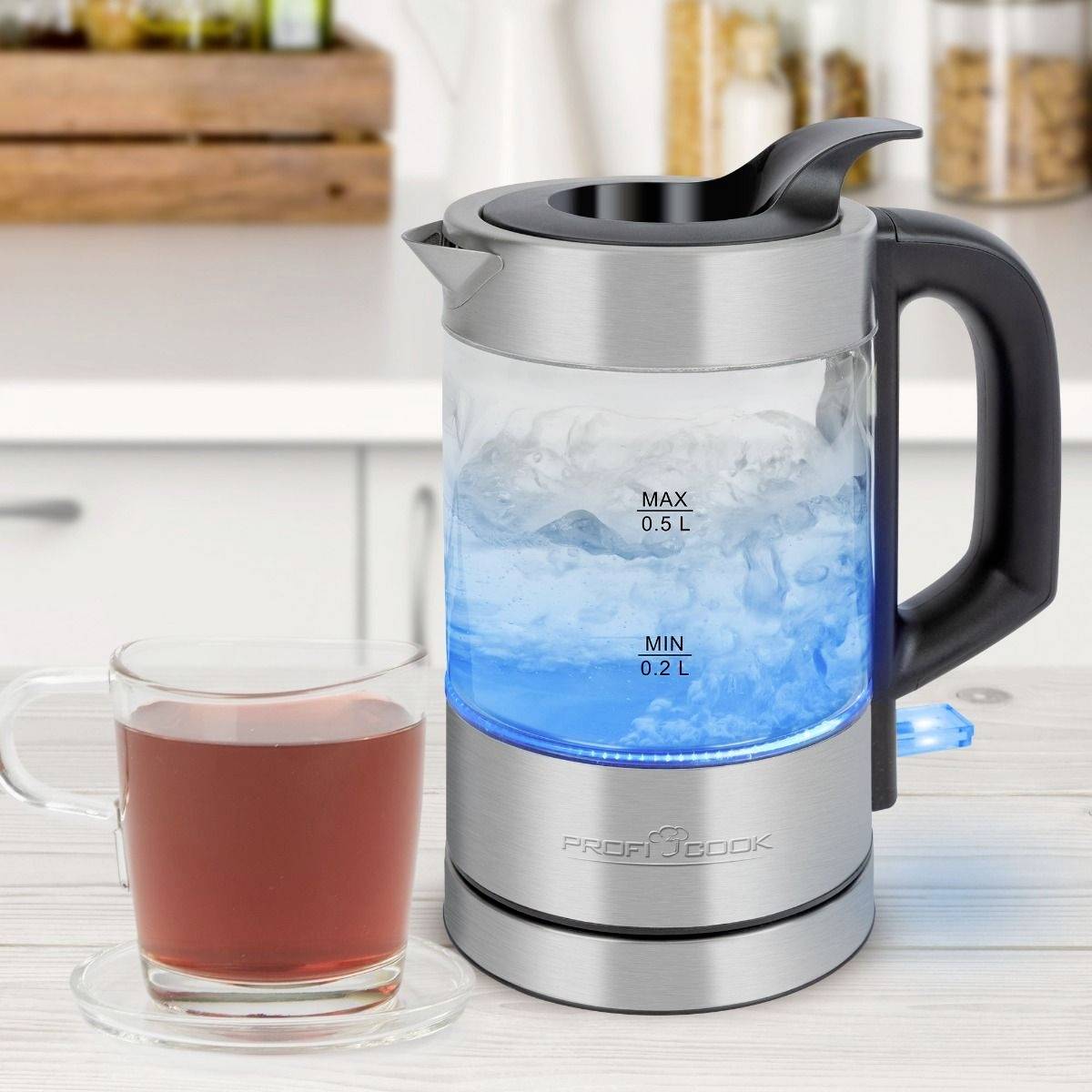 Ein Wasserkocher mit blauer LED-Beleuchtung, zeigt das kochende Wasser. Daneben steht eine Glas-Tasse mit Tee auf einer Küchenarbeitsplatte.