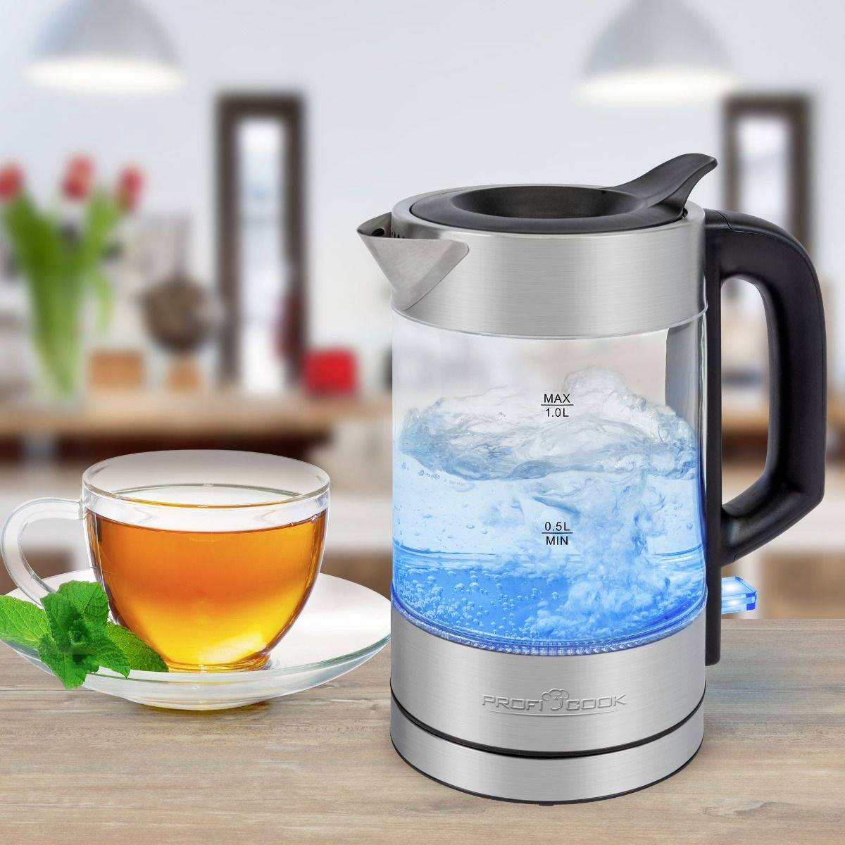 Ein Glaswasserkocher mit blauer LED-Beleuchtung neben einer Tasse Tee mit Minze auf einem Tisch, im Hintergrund eine verschwommene Küche.
