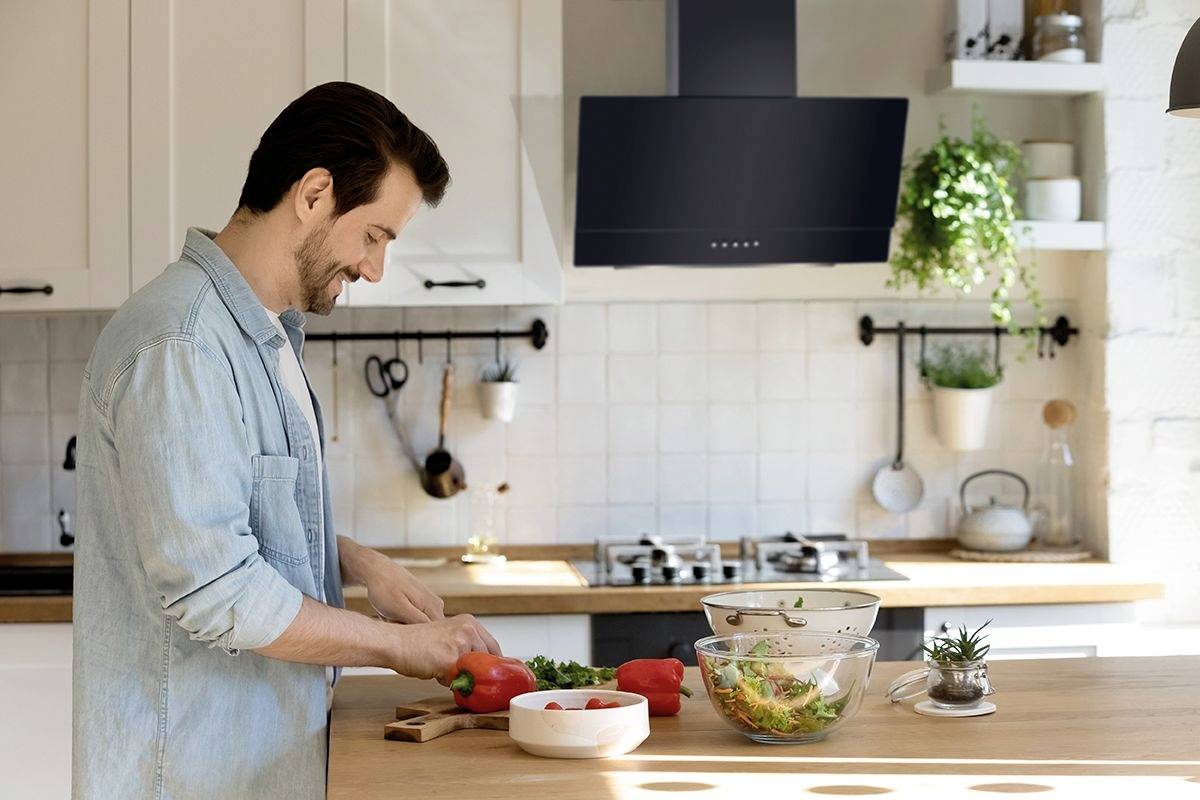 Ein Mann schneidet Gemüse in einer modernen Küche. Auf der Arbeitsplatte stehen Schüsseln mit Salatzutaten, und im Hintergrund sind Küchengeräte sichtbar.