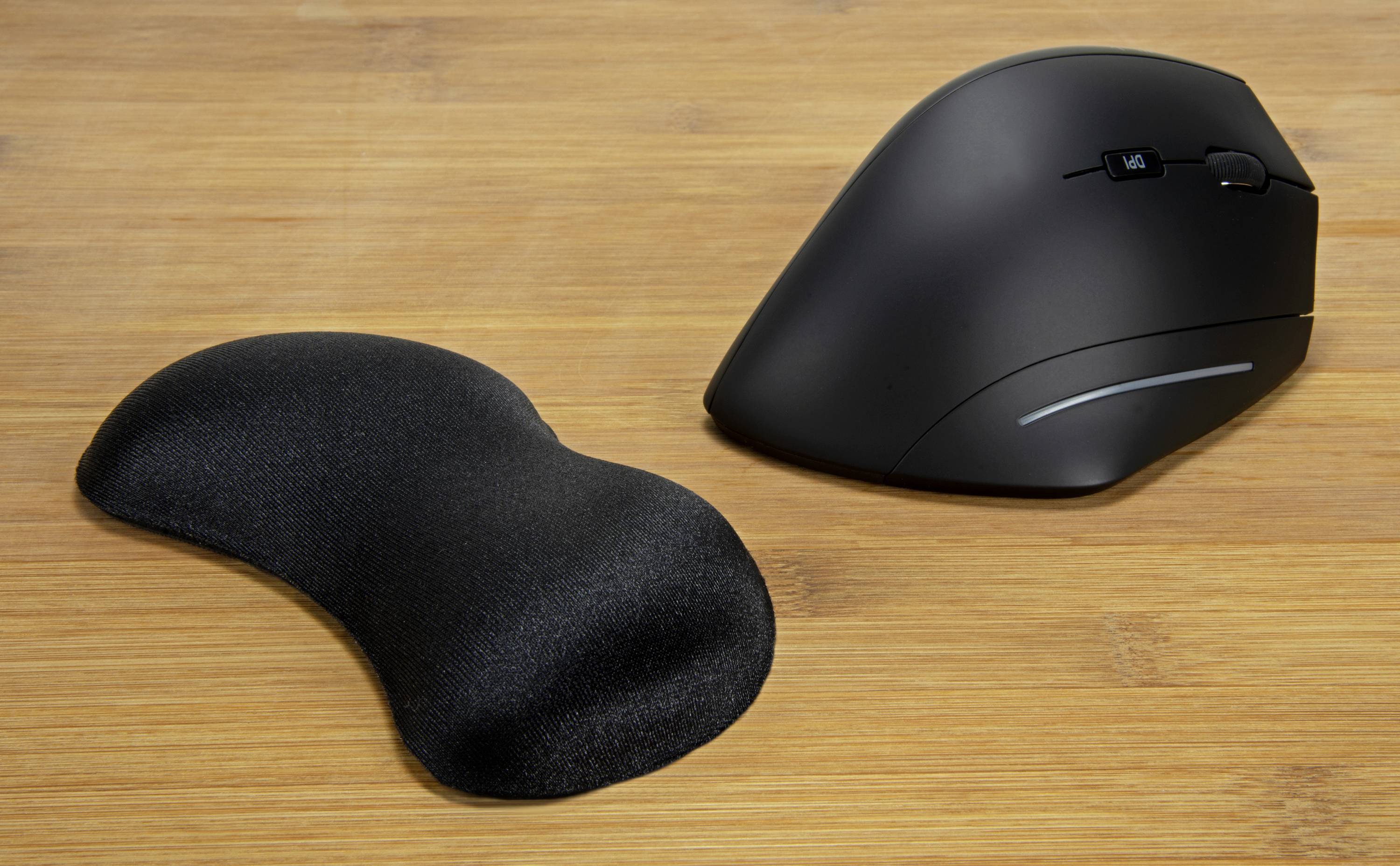 Eine schwarze ergonomische Computermaus mit anpassbarem Seitengriff liegt auf einem Holztisch neben einer Handgelenkauflage.