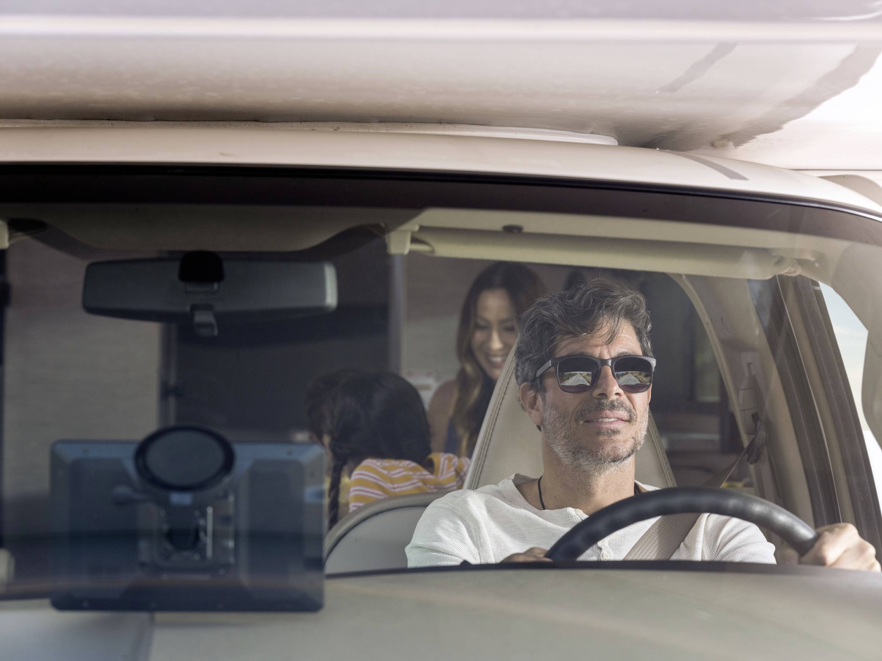 Ein Mann fährt ein Auto, während eine Frau und ein Kind im Hintergrund sitzen. Sie lächeln und genießen die gemeinsame Autofahrt.