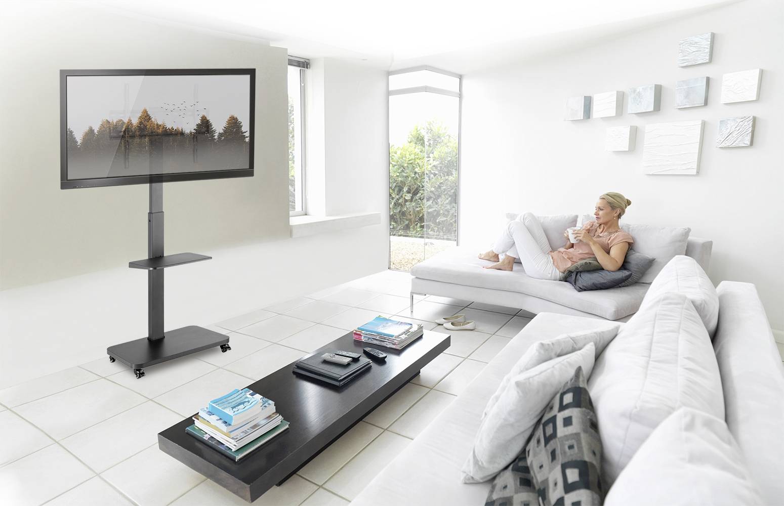 Frau sitzt auf einem Sofa in einem hellen Wohnzimmer, trinkt Kaffee und schaut einen Fernseher an, der auf einem Ständer steht.