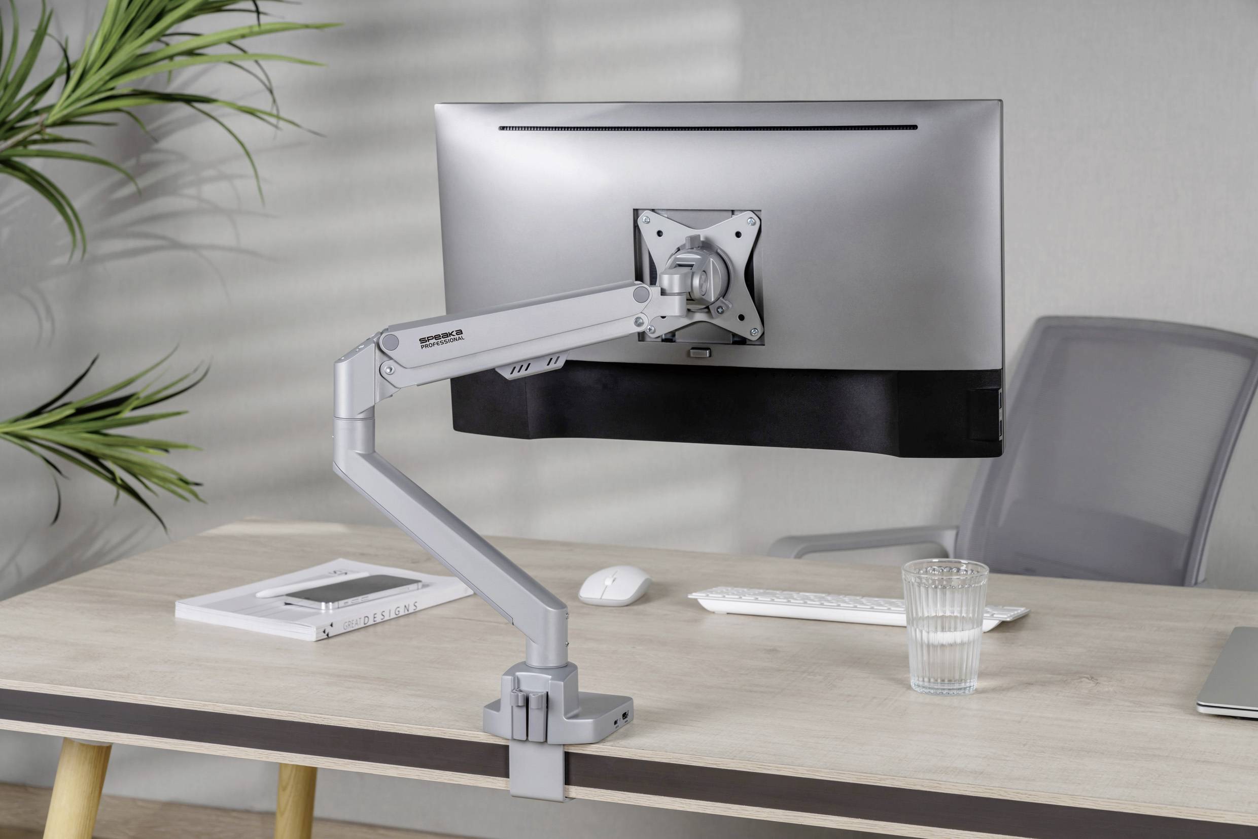 Ein Schreibtisch mit einem Computerbildschirm an einem verstellbaren Arm, Tastatur und Glas Wasser darauf; im Hintergrund eine graue Wand und Pflanze.