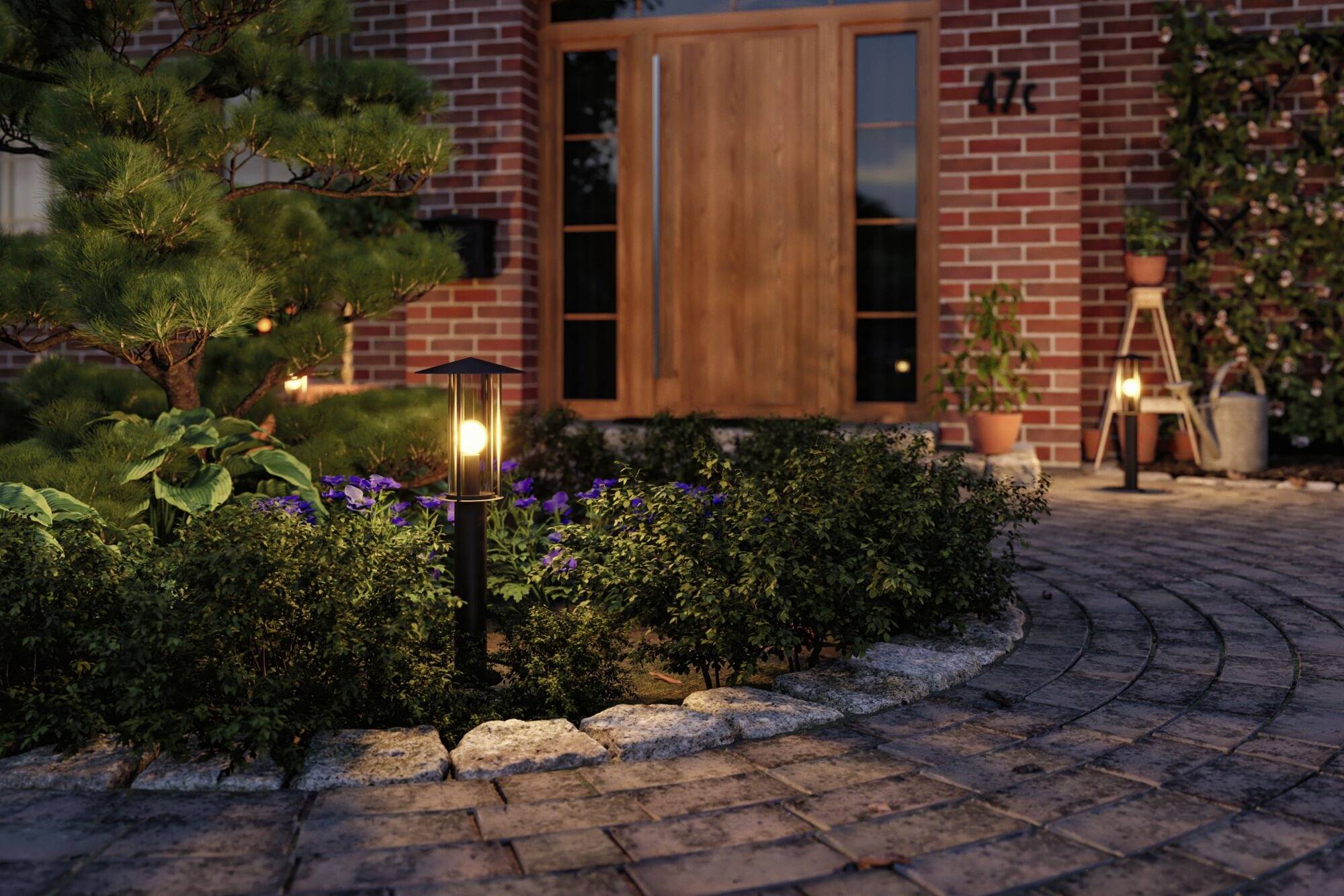 Ziegelstein-Hausfront bei Dämmerung, beleuchtet durch Gartenlampen. Ein Holzportal und Pflanzen schaffen eine einladende Atmosphäre.
