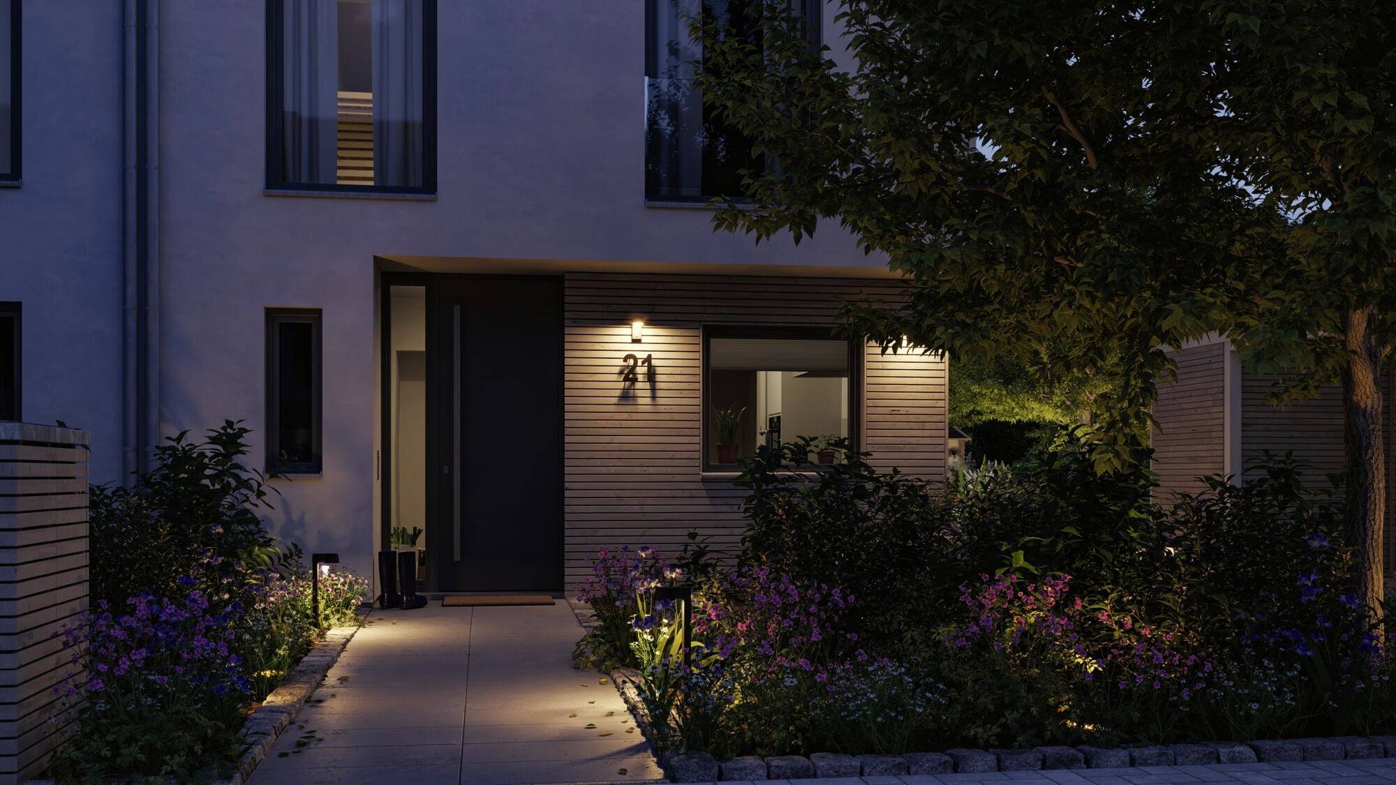 Zwei-stöckiges Haus bei Nacht mit beleuchtetem Eingang, umgeben von Gartenpflanzen und einem gepflasterten Weg.