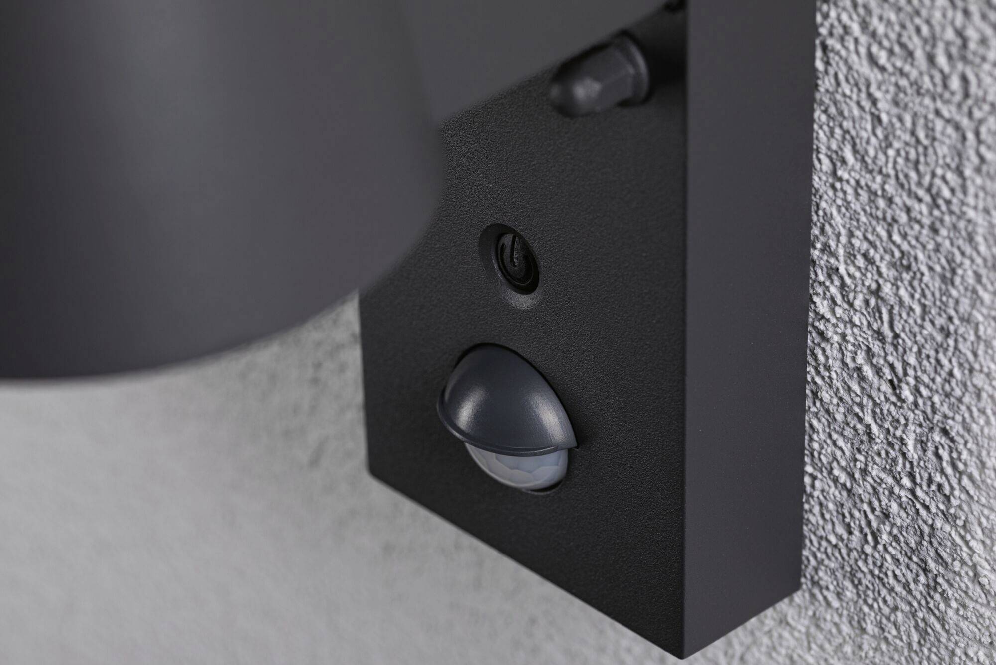 Bewegungsmelder an einer grauen Wand, nah aufgenommen. Hauptmerkmal ist der halbkreisförmige Sensor unter einem Schalter.