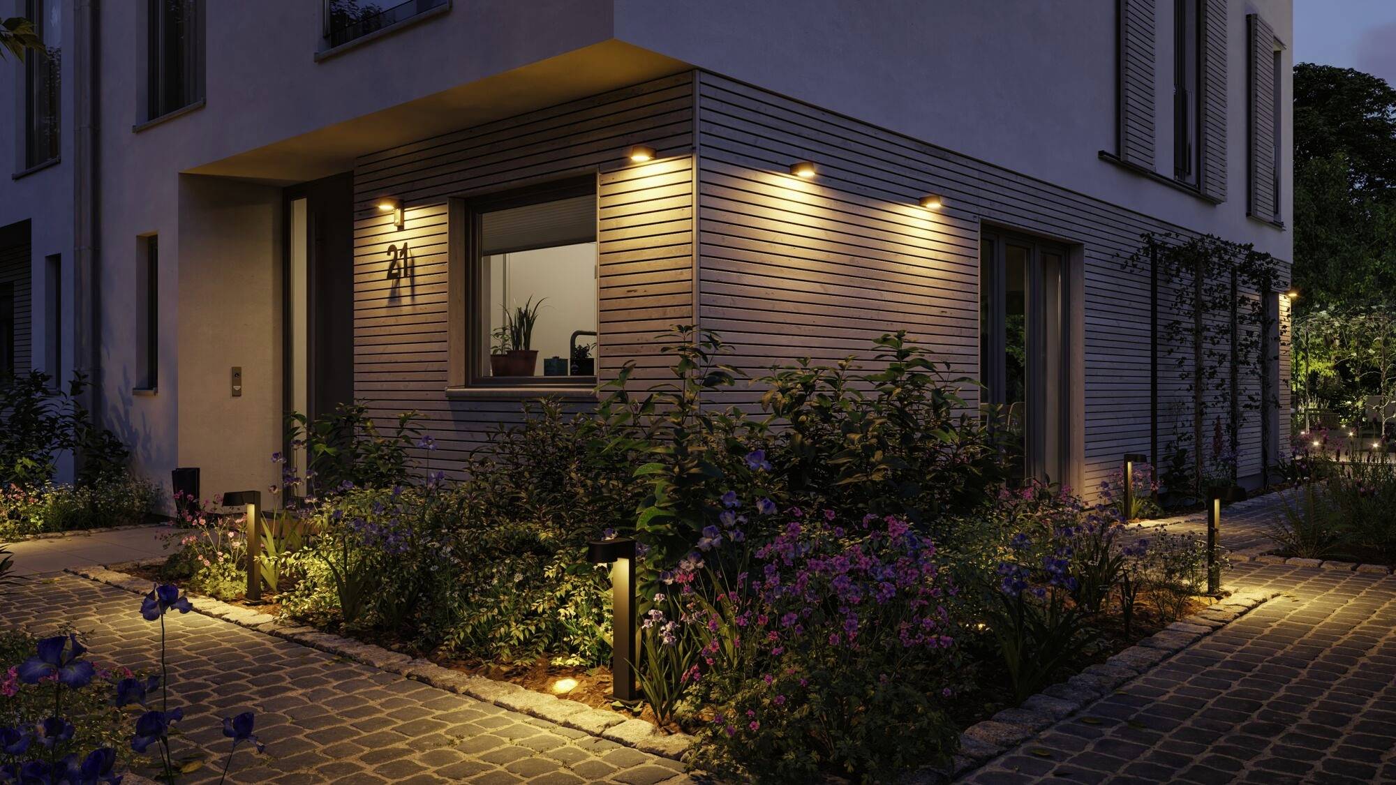 Eckansicht eines modernen Hauses bei Dämmerung, beleuchtet mit warmem Licht. Ein gepflegter Garten voller bunter Blumen umrahmt das Gebäude.