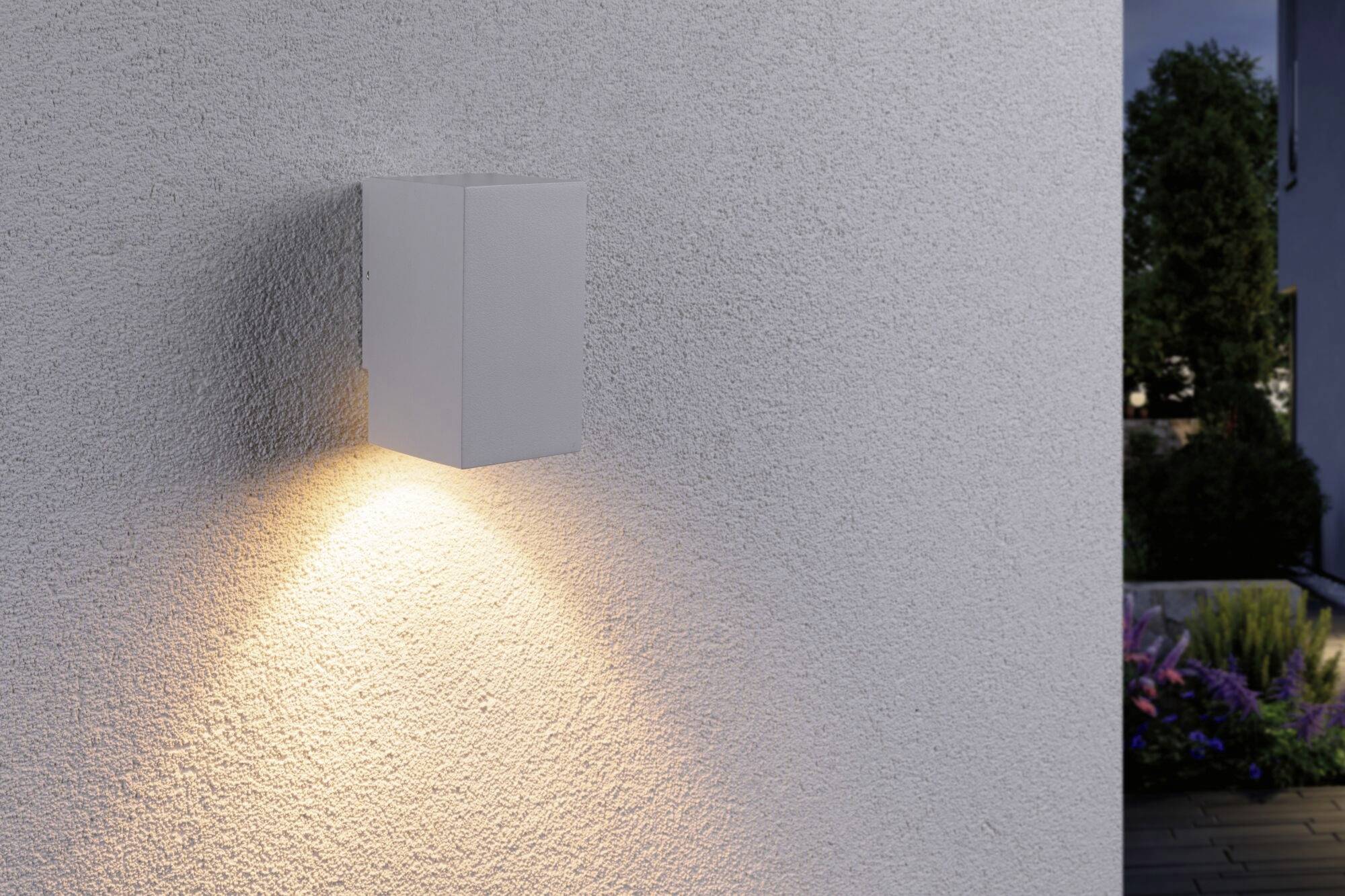 Außenwandlampe an einer hellen Wand, strahlt warmes Licht nach unten ab. Im Hintergrund sind Pflanzen und ein Haus erkennbar.