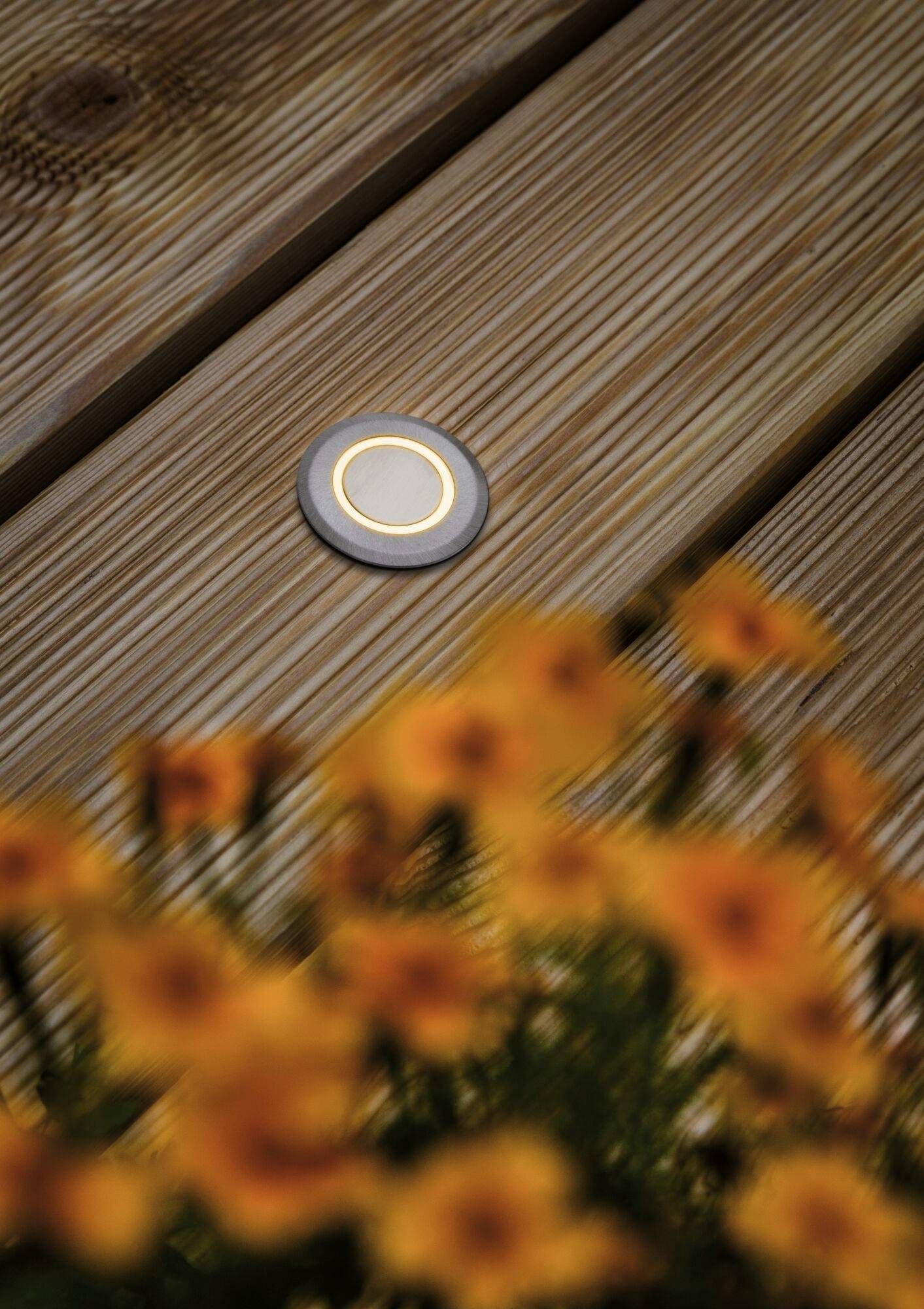 Eine kleine runde Bodenlampe ist in Holzplanken eingebettet. Im Vordergrund sind unscharfe, gelbe Blumen zu sehen.
