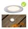 Runde LED-Leuchte in Holzterrasse eingebaut, mit weichem gelbem Licht, unter einem grünen Logo mit stilisiertem Glühwürmchen.
