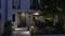 Eingang eines modernen Hauses bei Nacht, beleuchtet von mehreren Gartenlichtern und umgeben von Pflanzen. Hausnummer 21 sichtbar links der Tür.