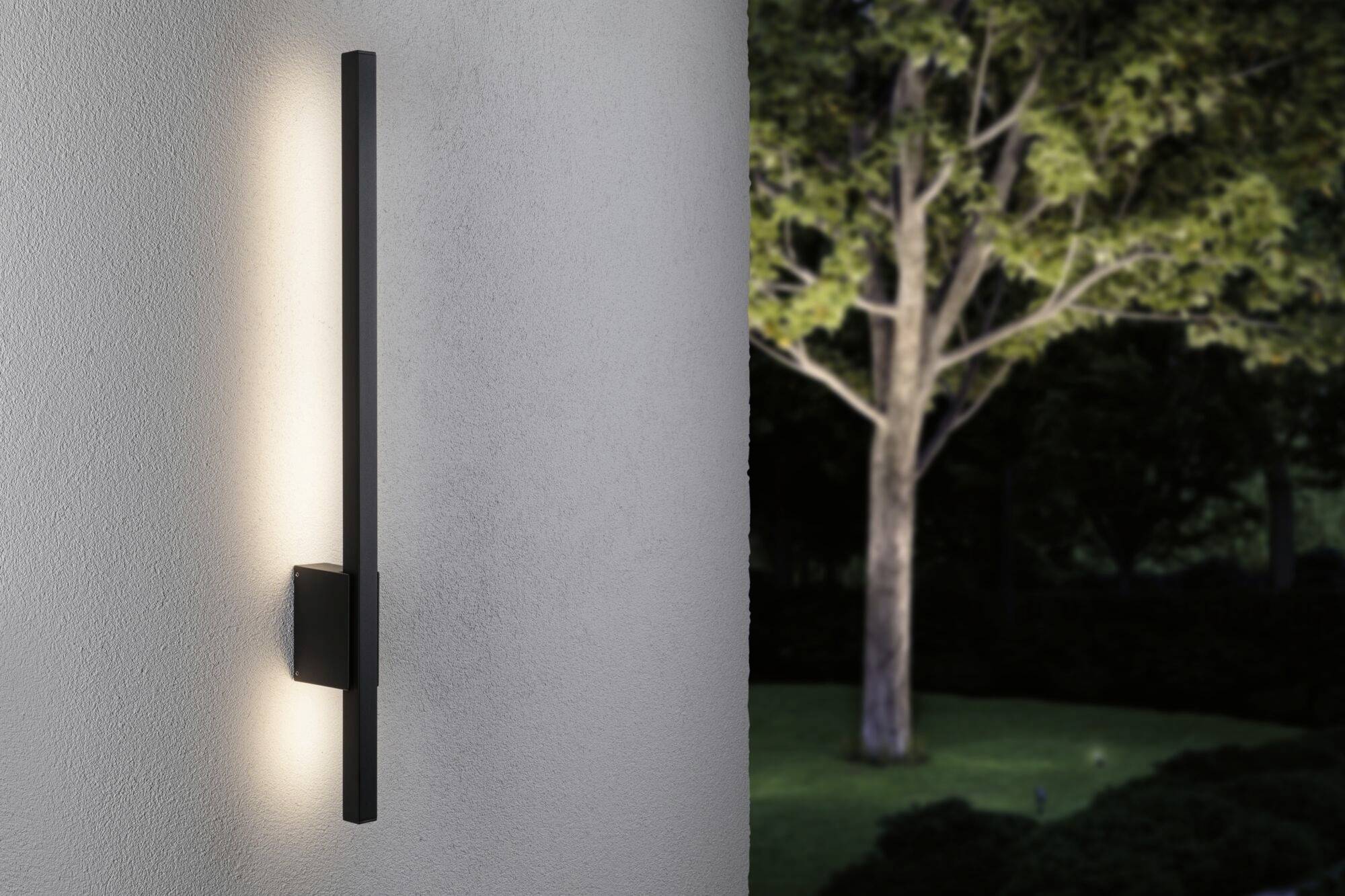 Eine schlanke schwarze Außenwandleuchte leuchtet an einer weißen Wand, mit einem beleuchteten Baum und Garten im Hintergrund bei Nacht.