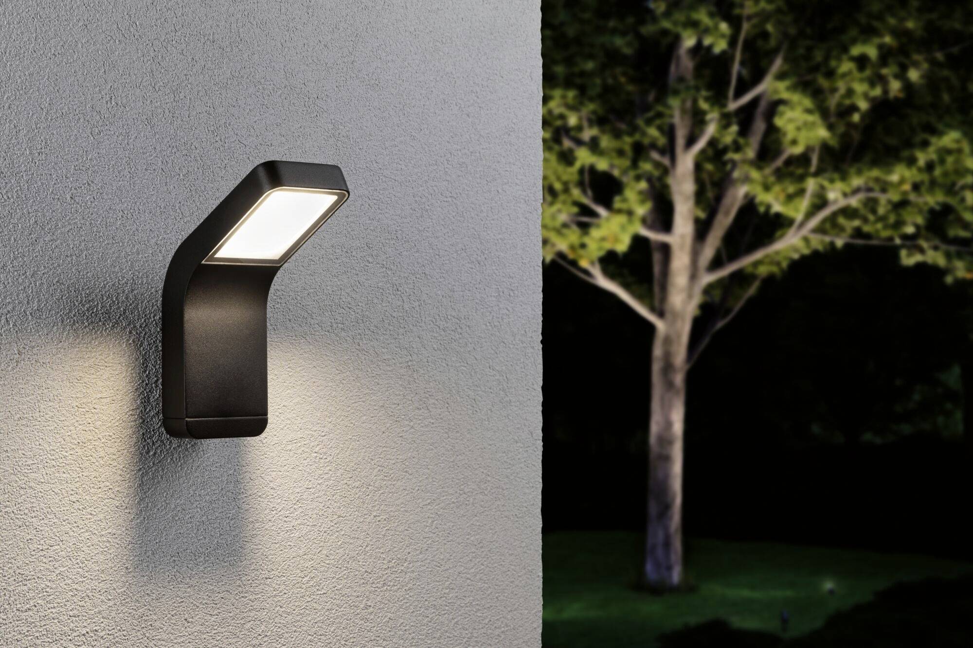 Moderne Außenwandleuchte an grauer Wand, beleuchtet in der Nacht. Im Hintergrund ist ein Baum in einem Garten zu sehen.