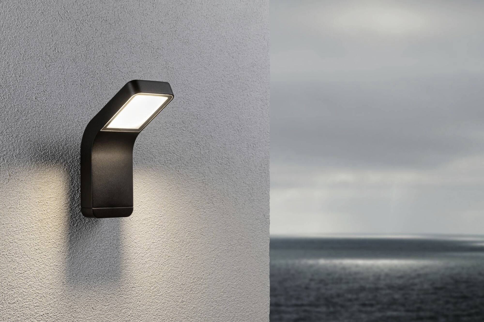 Die Wandlampe leuchtet an einer grauen Wand. Im Hintergrund ist ein ruhiges Meer unter einem bewölkten Himmel sichtbar.
