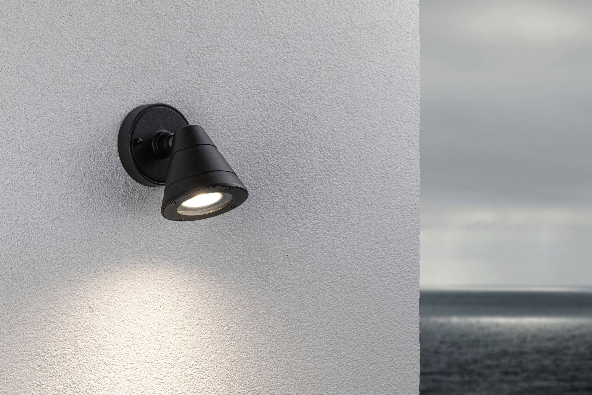 Schwarz-graue Außenleuchte an einer weißen Hauswand. Sie beleuchtet subtil den Wandbereich. Im Hintergrund ist das Meer sichtbar.