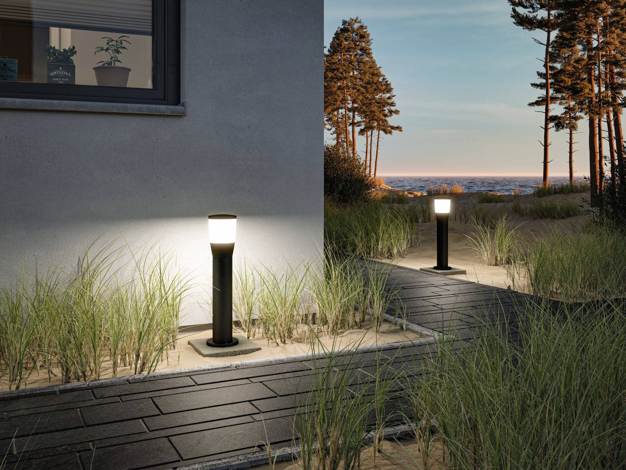 Ein beleuchteter Gartenweg bei Dämmerung mit zwei schwarzen Gartenlampen, umgeben von Sand und Gras, führt zu einer Küstensicht.