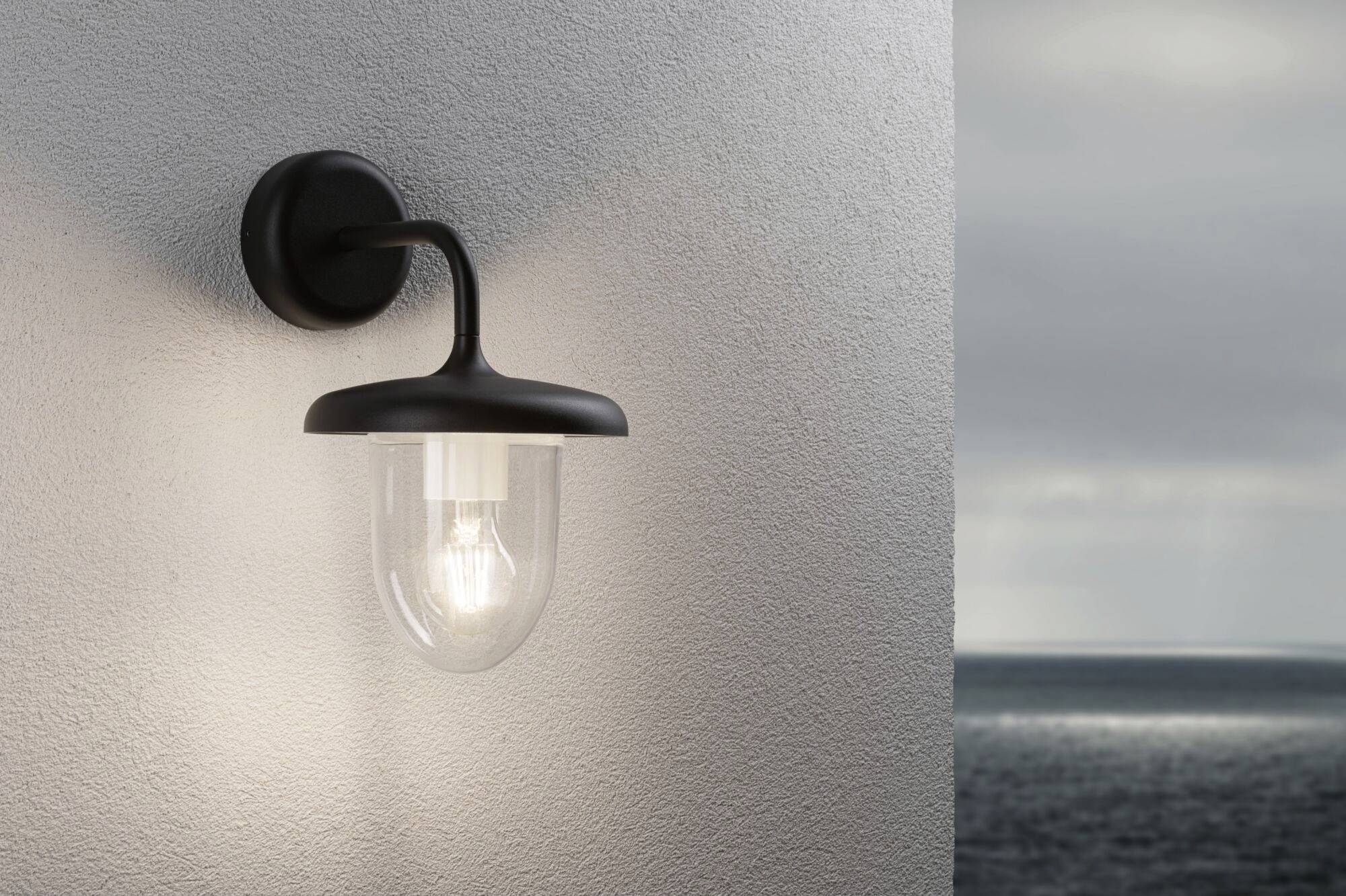 Wandleuchte mit Glasabdeckung an einer Wand, beleuchtet. Im Hintergrund ist ein bewölkter Himmel über dem Meer sichtbar.