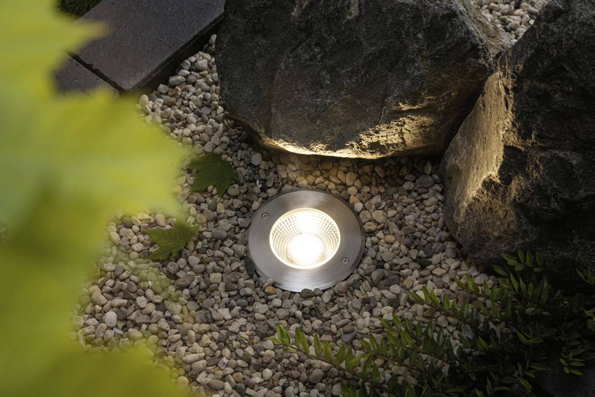 Bodenleuchte zwischen Steinen und Pflanzen in einem Garten, die eine warme Lichtatmosphäre schafft.