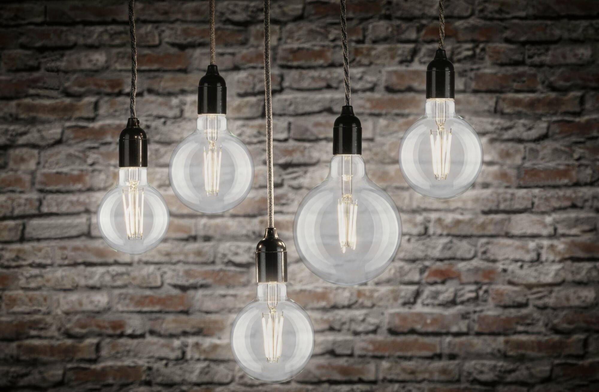Fünf hängende Glühbirnen mit sichtbaren Filamenten vor einer Ziegelwand, unterschiedlich große Birnen.