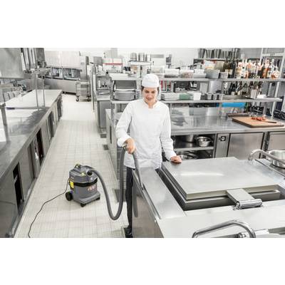 Ein Koch in einer gewerblichen Küche verwendet einen Staubsauger, um Edelstahloberflächen zu reinigen, umgeben von industriellen Geräten und Kochgeschirr.