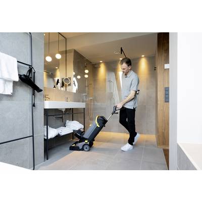 Ein Mann benutzt eine Bodenreinigungsmaschine in einem modernen Badezimmer mit Dusche, großen Spiegeln und Handtüchern auf dem Regal.