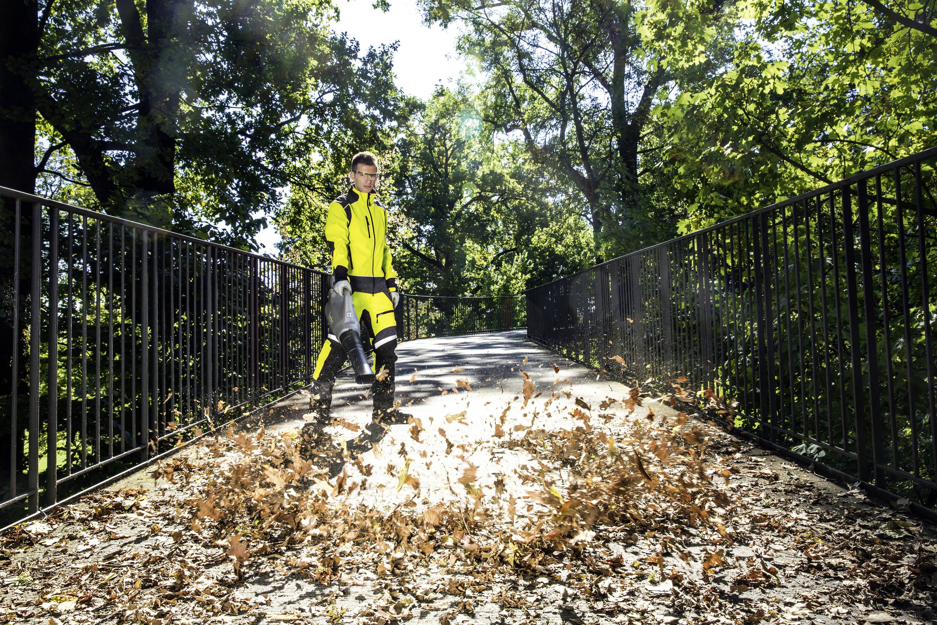 Eine Person in gelber Schutzkleidung bläst Blätter mit einem Laubbläser auf einer Brücke in einem sonnigen Park.