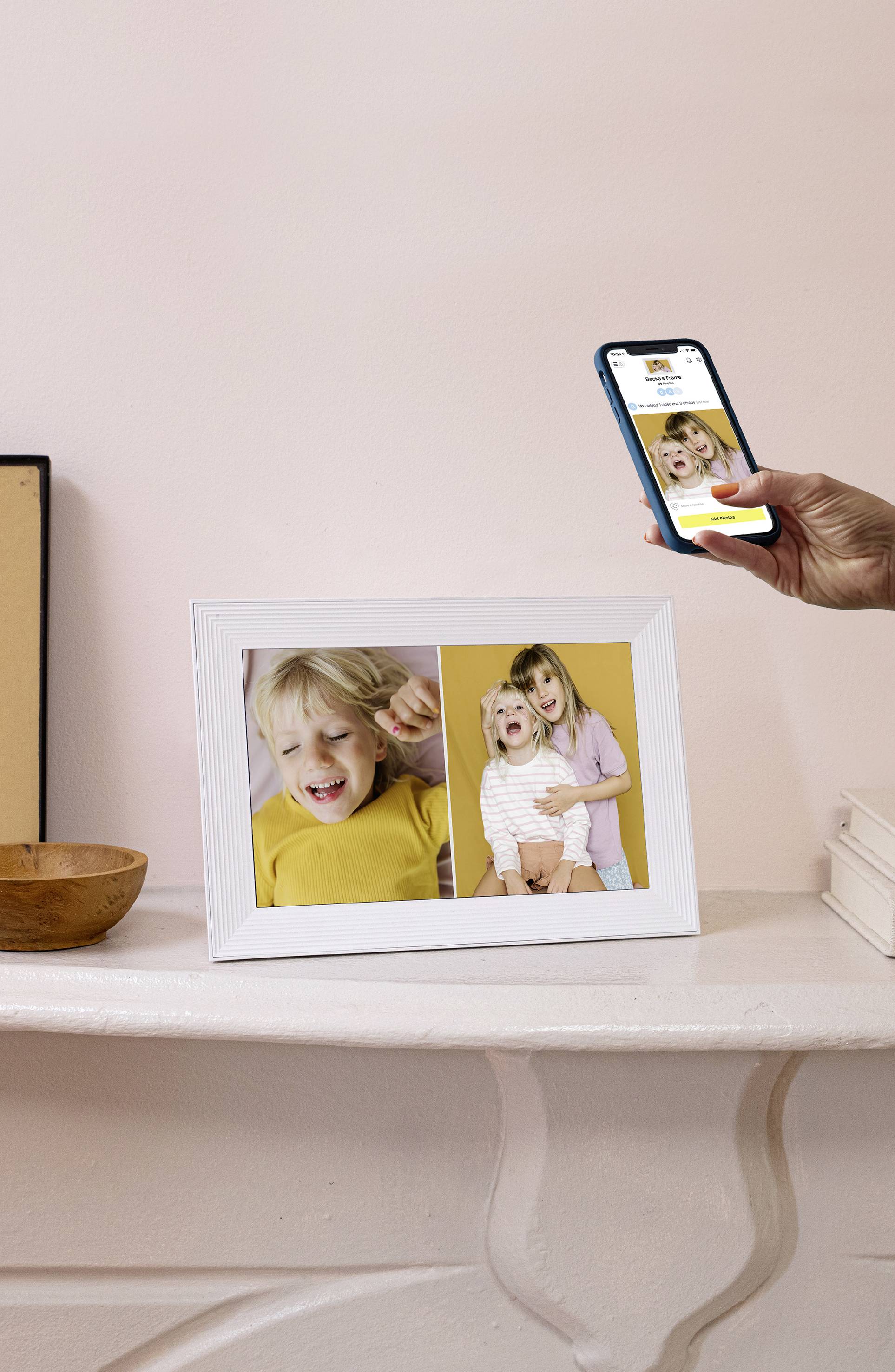 Ein gerahmtes Foto auf einem Regal zeigt zwei Kinder. Eine lächelnde Hand hält ein Smartphone, das dasselbe Bild auf dem Bildschirm zeigt.