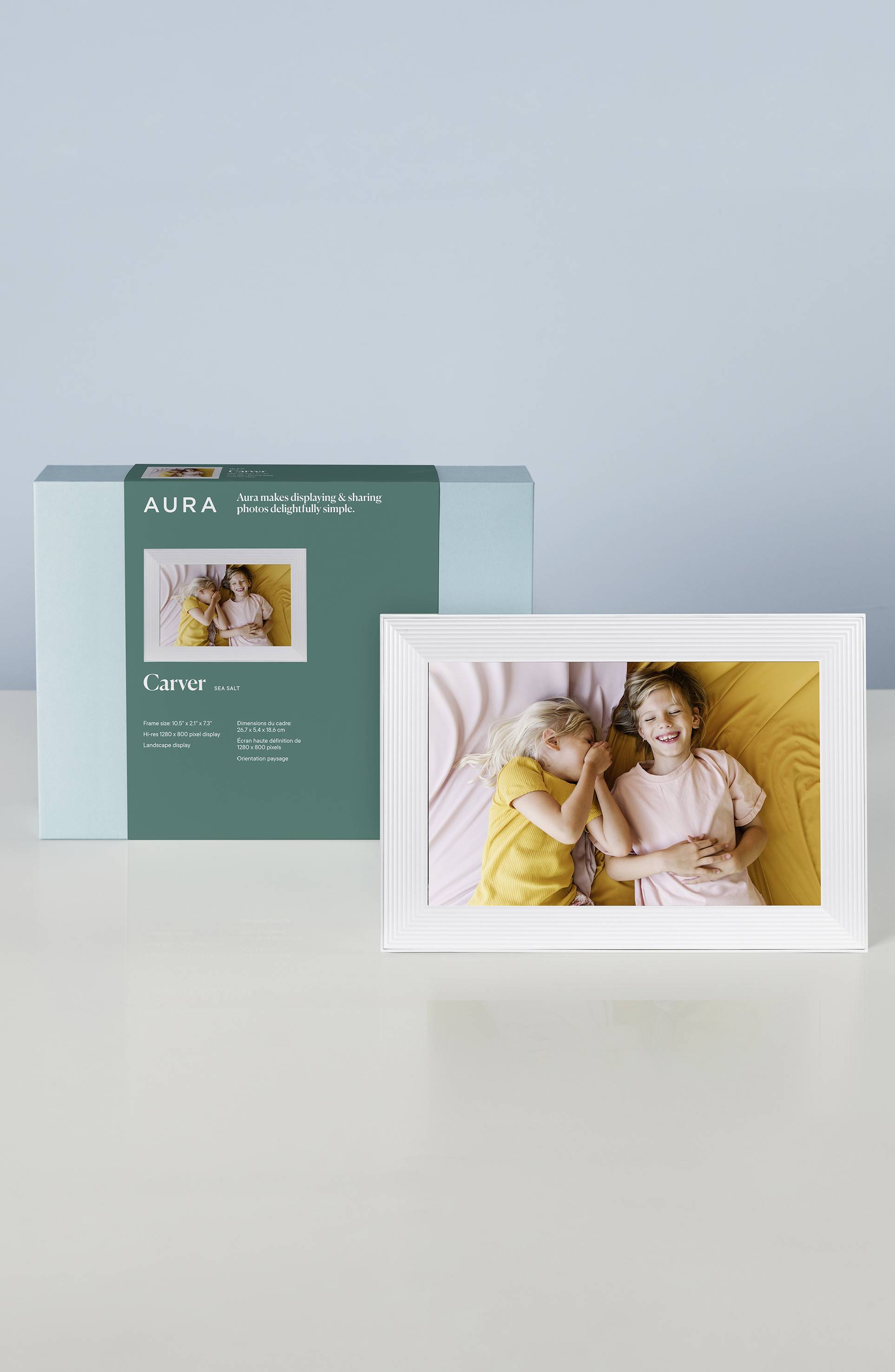 Digitaler Bilderrahmen auf einem Tisch, zeigt Foto von zwei lachenden Kindern im gelben und rosa Hemd. Verpackung im Hintergrund.