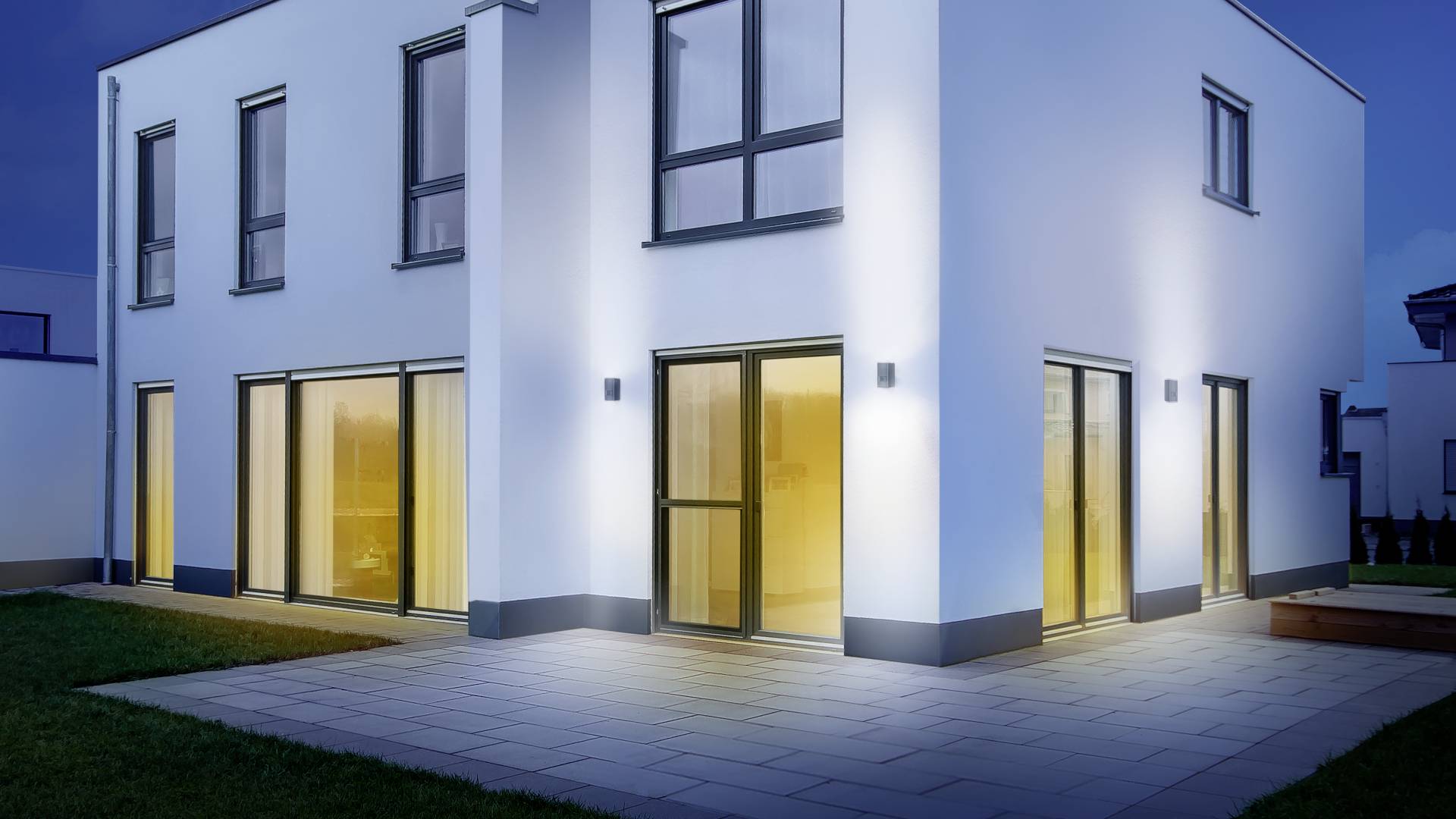 Modernes zweistöckiges Haus bei Nacht mit großen Fenstern, die warmes Licht ausstrahlen. Außenbeleuchtung hebt die Fassade hervor.