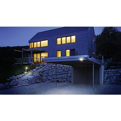 Ein modernes Haus mit großen, erleuchteten Fenstern bei Nacht, umgeben von einem Garten und Steinmauern, mit Carport im Vordergrund.