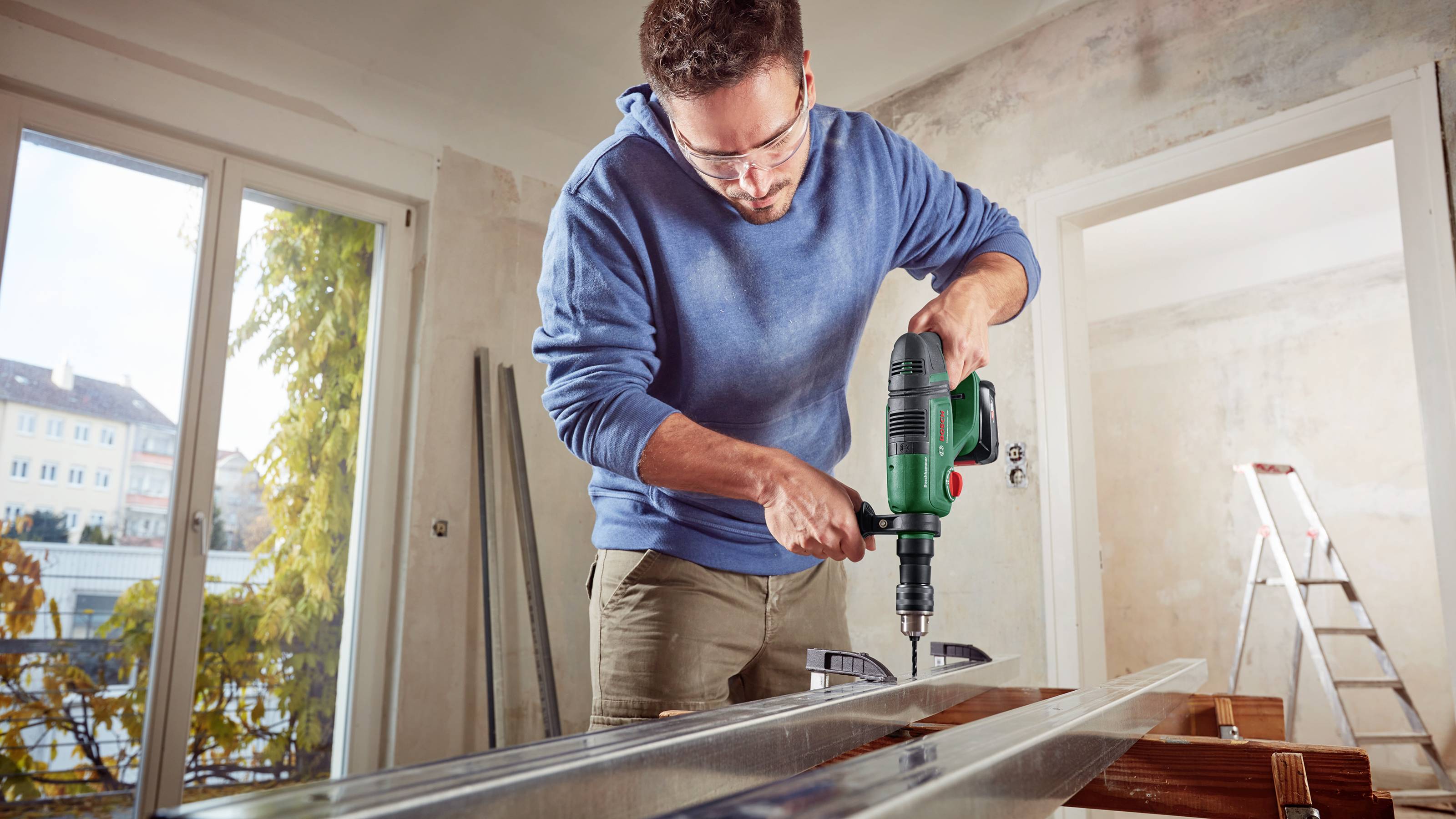 Ein Mann in Schutzbrille und blauem Hoodie bohrt mit einer elektrischen Bohrmaschine in einem Raum, der renoviert wird. Im Hintergrund ist eine Leiter.