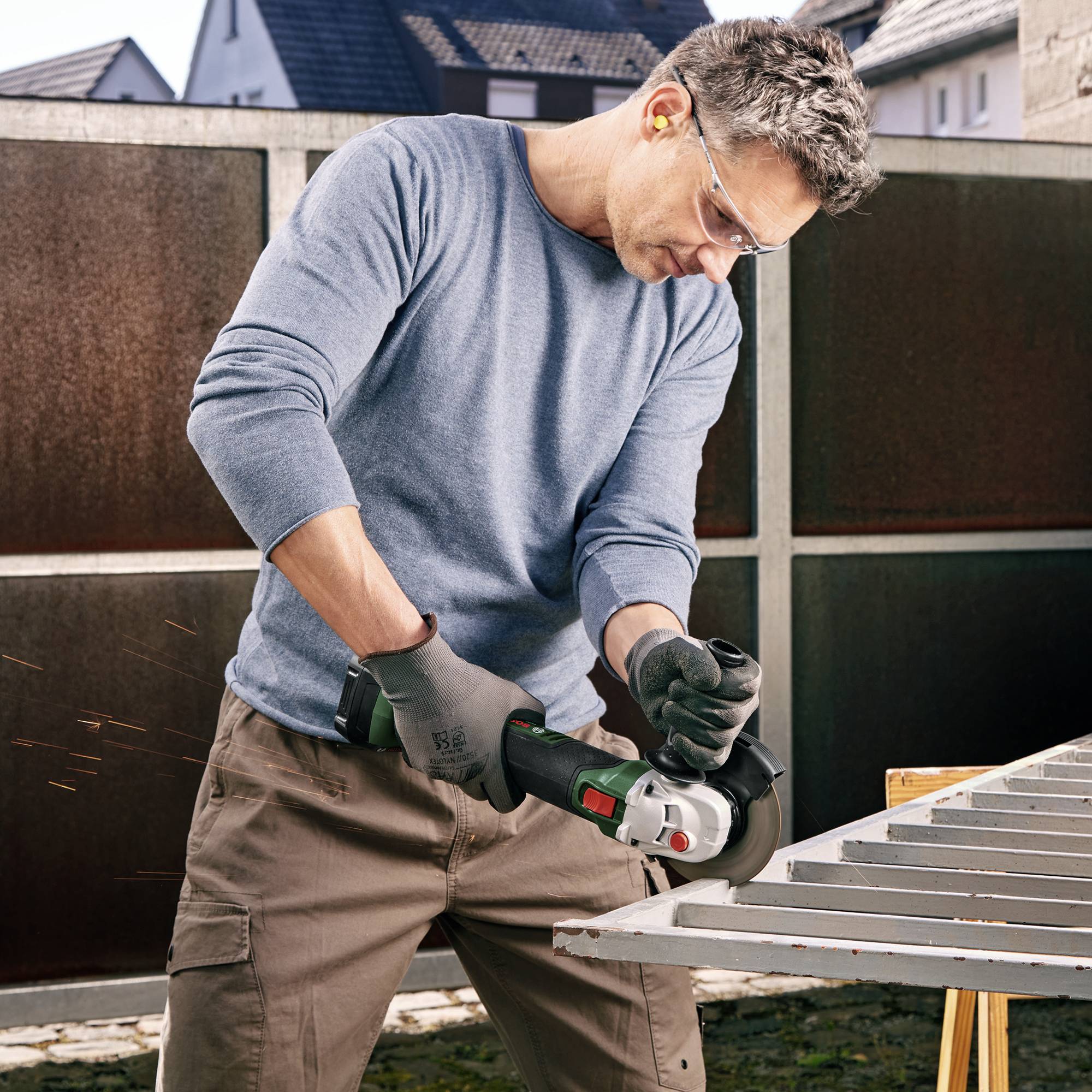 Ein Mann in Schutzbrille und Ohrschützern nutzt einen Winkelschleifer, um Metallstäbe zu schneiden. Funken fliegen während der Arbeit.