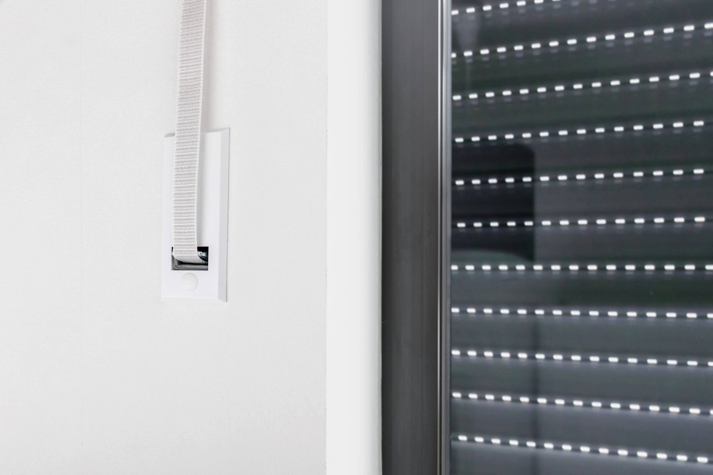 Jalousie mit Gurt an der Wand neben einem Fenster. Sie ist teilweise geschlossen, Licht dringt durch die Schlitze.