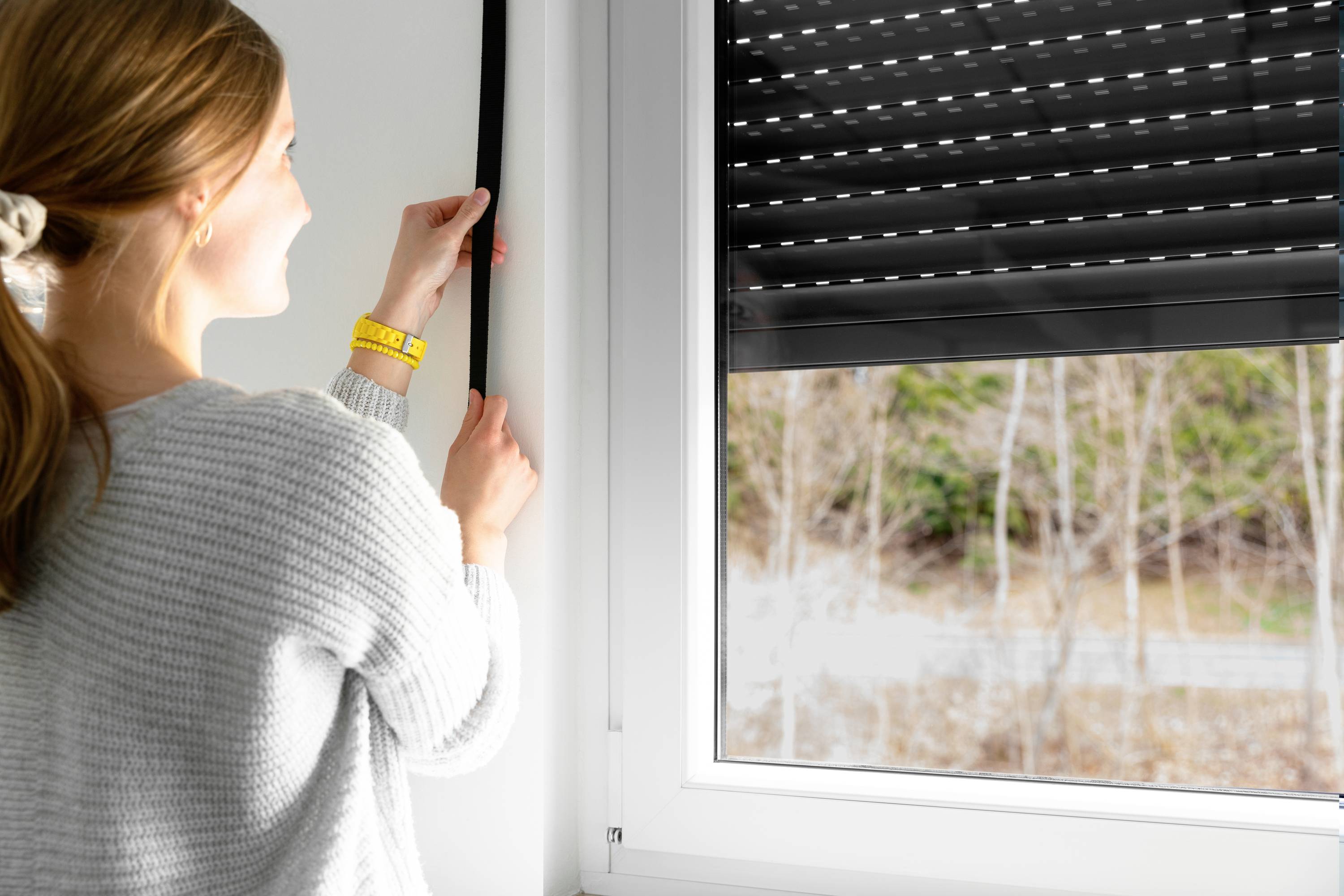 Eine Frau öffnet ein Fensterrollo, durch das sie nach draußen blickt. Im Hintergrund sind Bäume zu sehen.