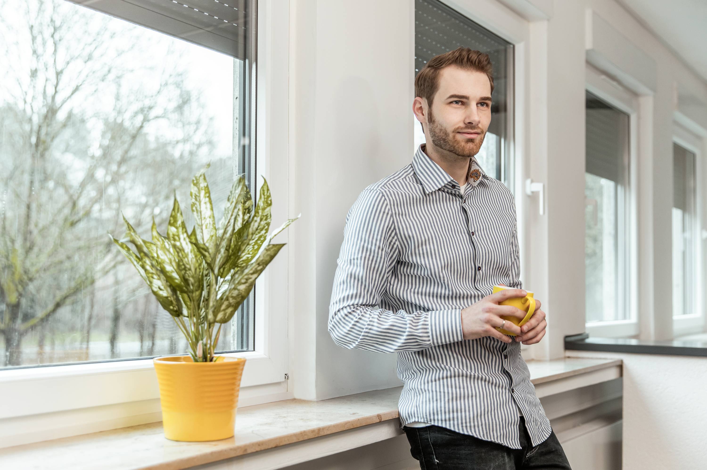 Ein Mann steht neben einem Fenster und hält eine gelbe Tasse. Neben ihm steht eine Pflanze in einem gelben Topf auf der Fensterbank.