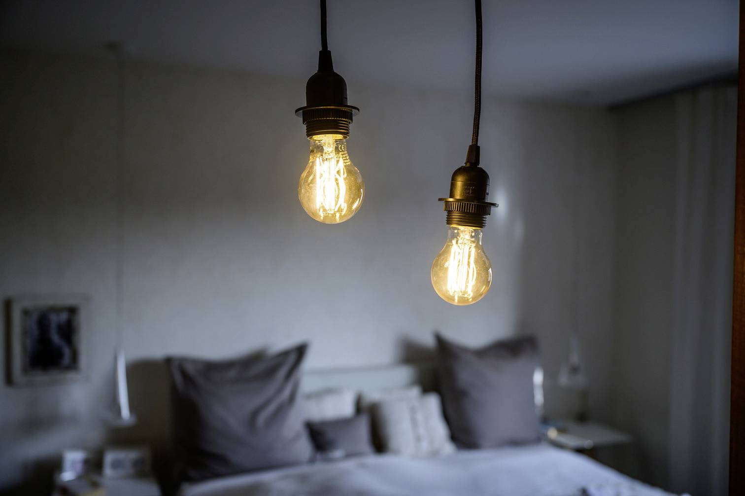 Zwei beleuchtete Glühbirnen hängen von der Decke in einem Schlafzimmer, im Hintergrund ist ein Bett mit grauen Kissen sichtbar.