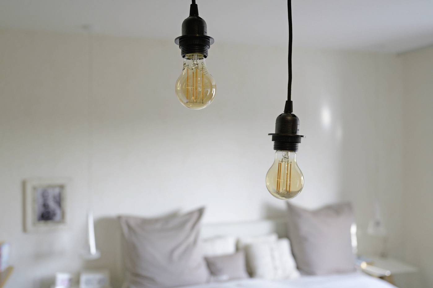 Zwei hängende Glühbirnen im Vordergrund, verschwommenes Schlafzimmer im Hintergrund mit Bett und Kissen.