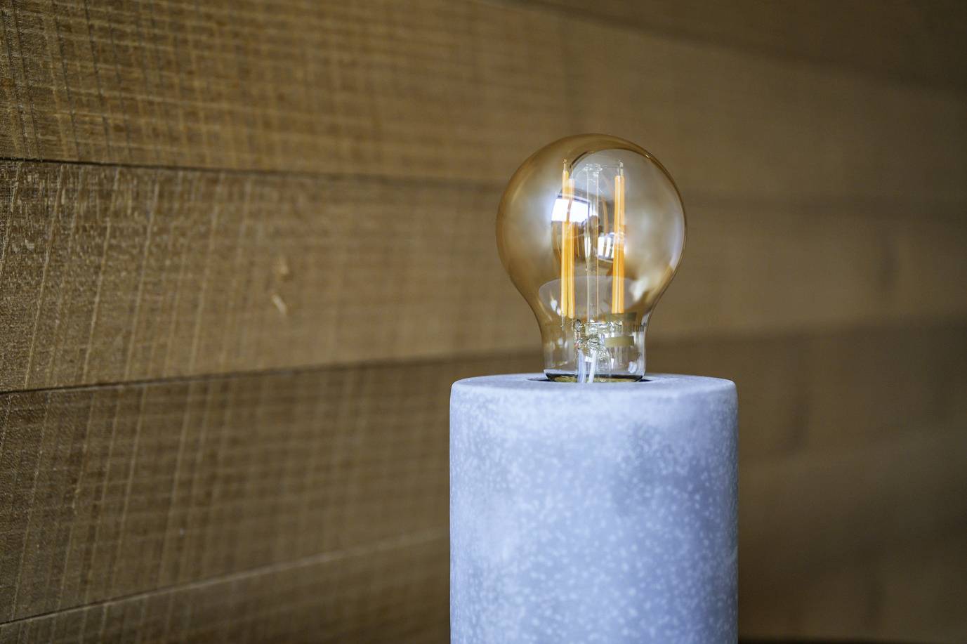 Eine Glühbirne auf einem grauen Betonständer vor einer Holzwand. Die Birne leuchtet, und der Hintergrund ist unscharf.