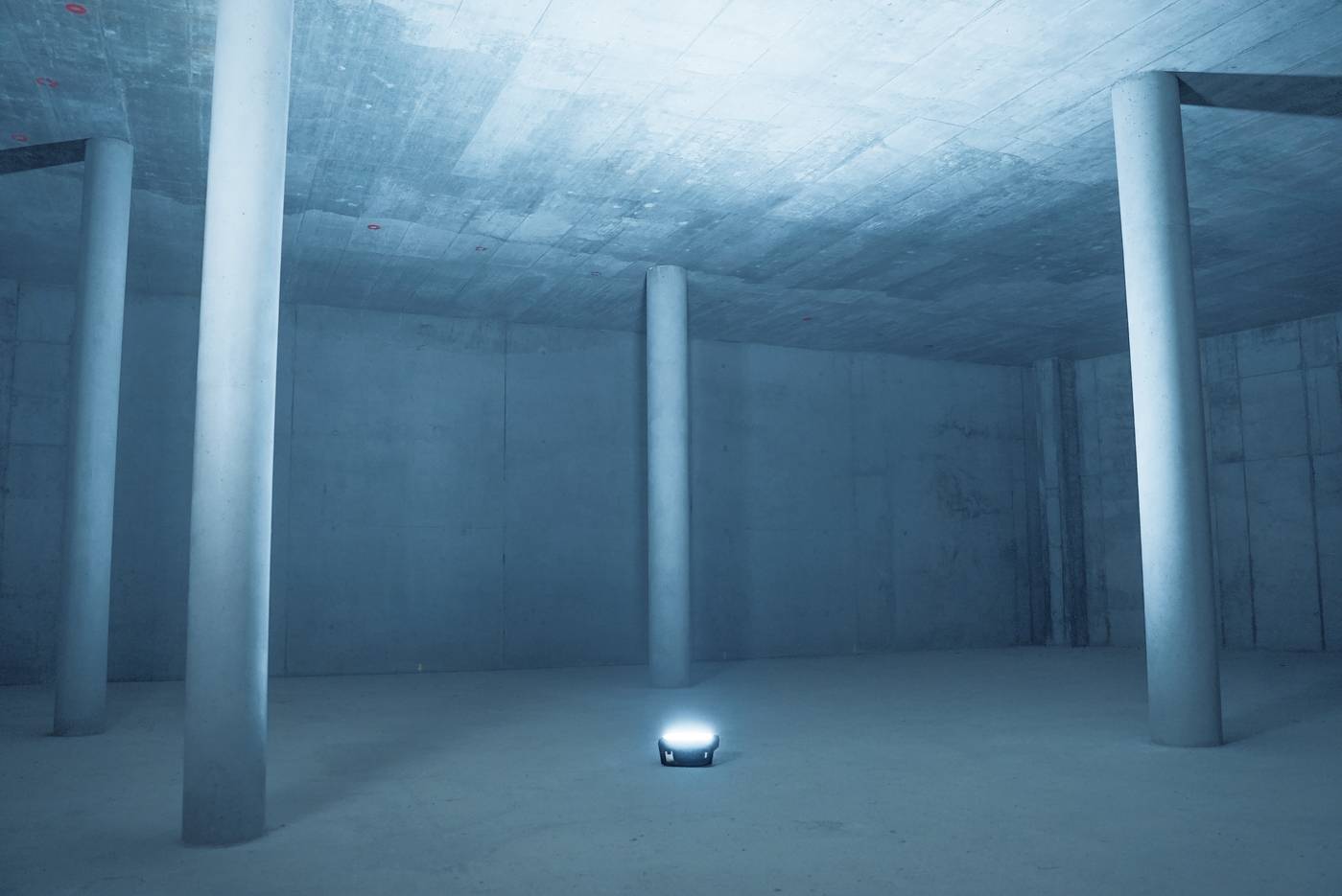 Ein leerer, blauer Betonraum mit hohen Säulen und einer leuchtenden Lampe in der Mitte.