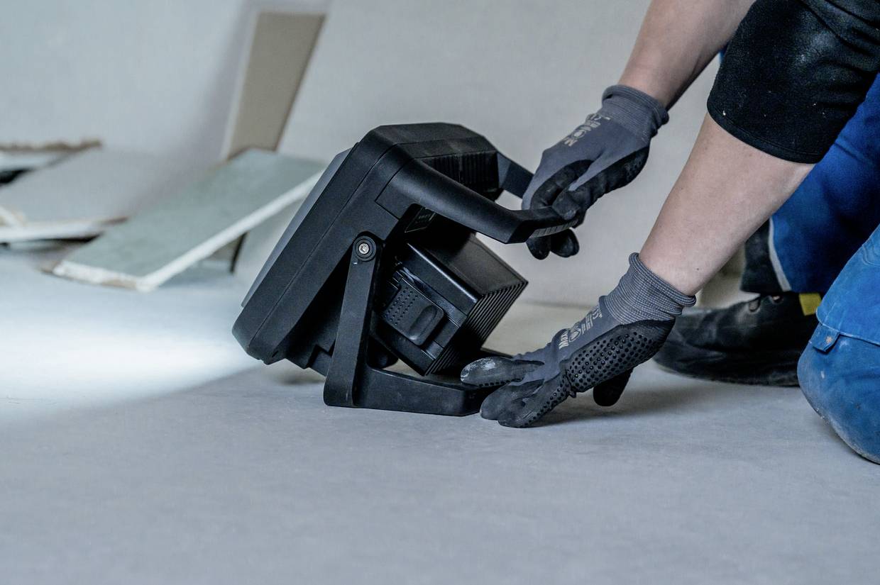 Eine Person in Arbeitshandschuhen richtet einen Baustrahler aus. Der Raum ist im Bau, mit Gipskartonplatten im Hintergrund.